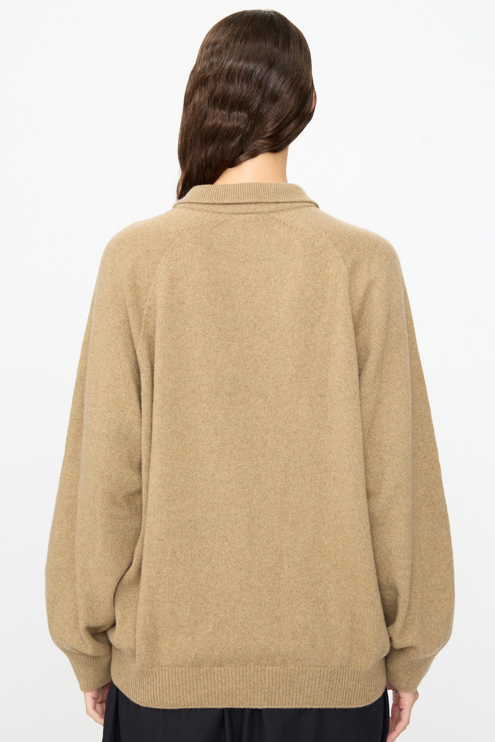 A person with long, wavy brown hair is shown from the back against a plain white background, wearing the Baserange Soar Polo in Rovere Brown—a loose-fitting cotton knit sweater—paired with a dark skirt.