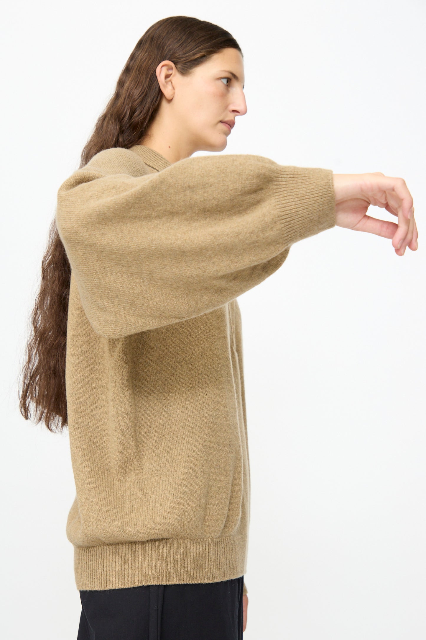 A person with long brown hair is wearing the Baserange Soar Polo in Rovere Brown, a loose cotton knit sweater with balloon sleeves, standing sideways with their right arm extended forward against a plain background.