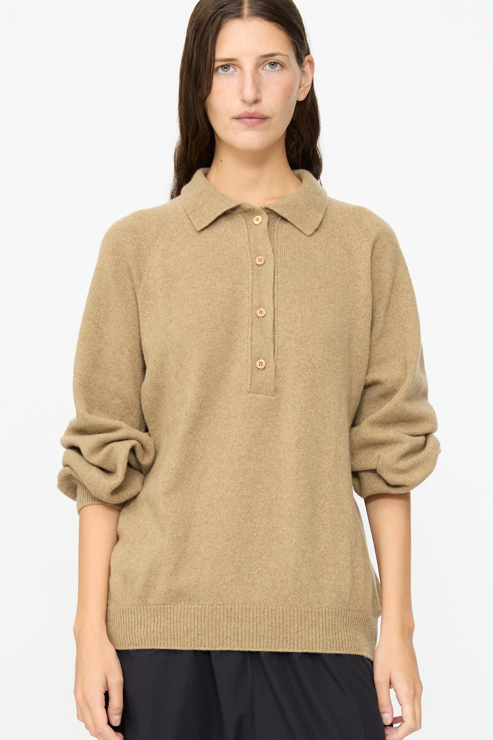 A person wearing the Baserange Soar Polo in Rovere Brown, a beige cotton knit long-sleeve sweater with a collar and buttons, stands against a plain white background.