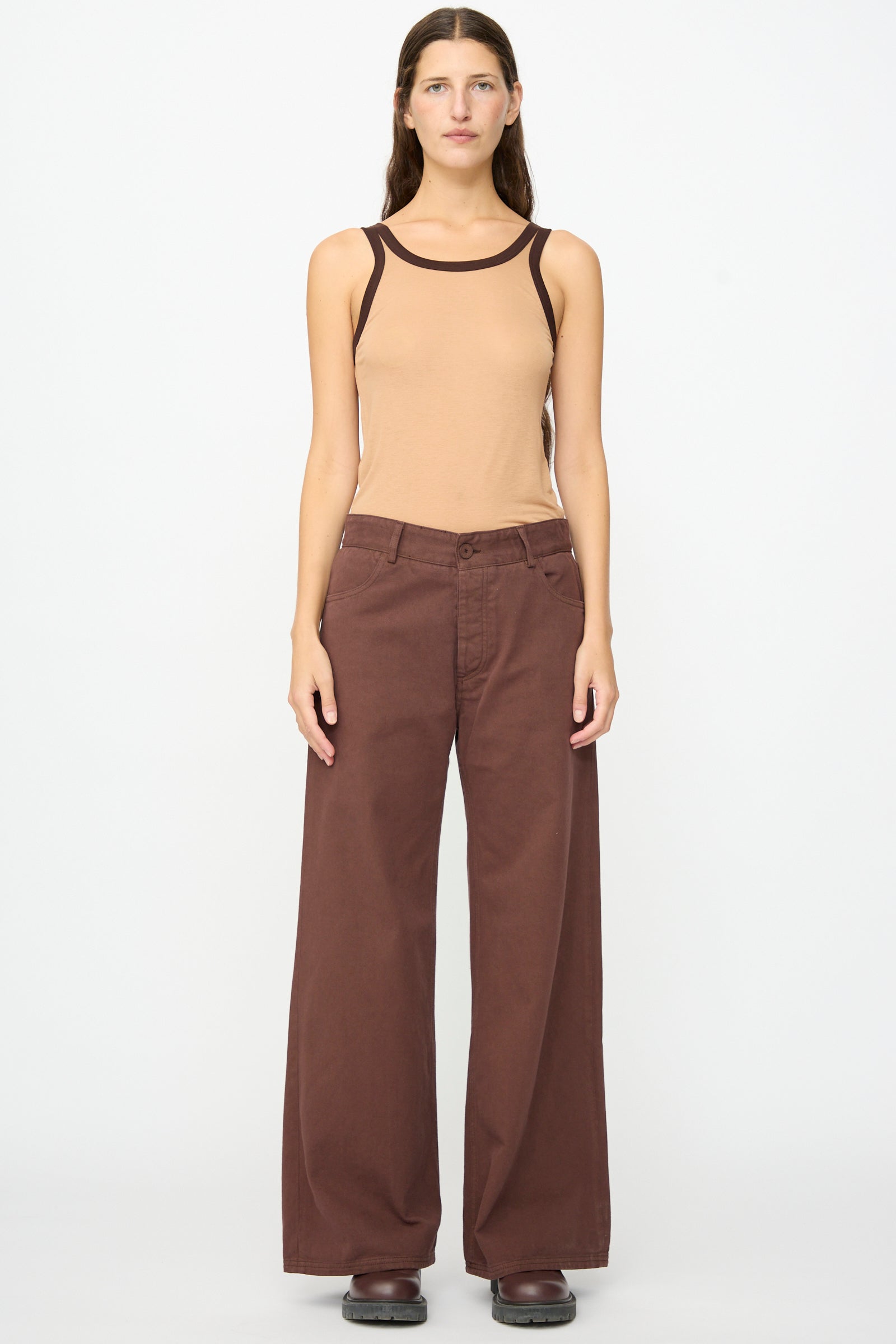 A person stands facing forward against a plain white background, wearing wide-legged Baserange Tajo Pant Wide in Cata Brown, paired with a tan sleeveless top with dark trim and dark shoes.
