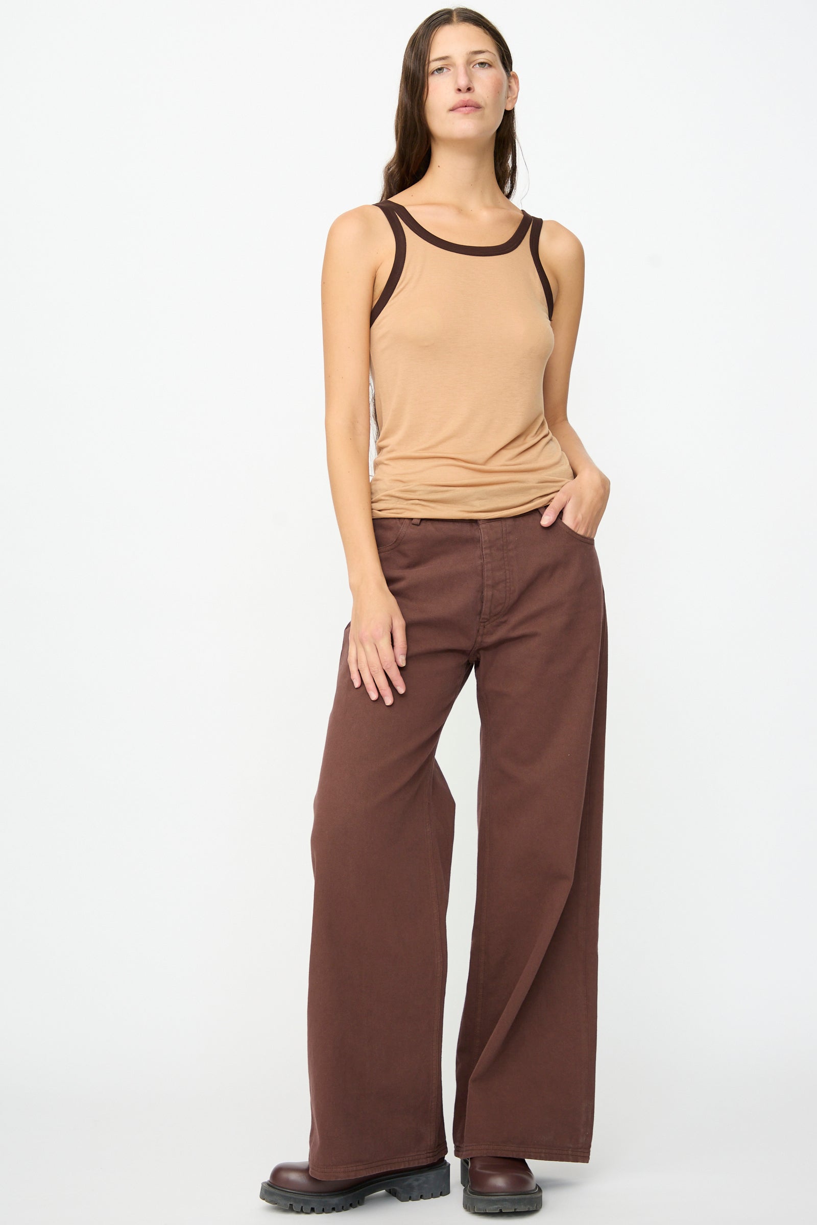 A woman stands against a white background wearing a tan tank top with dark trim, the Baserange Tajo Pant Wide in Cata Brown—wide-leg gabardine cotton pants made in Portugal—and brown shoes.