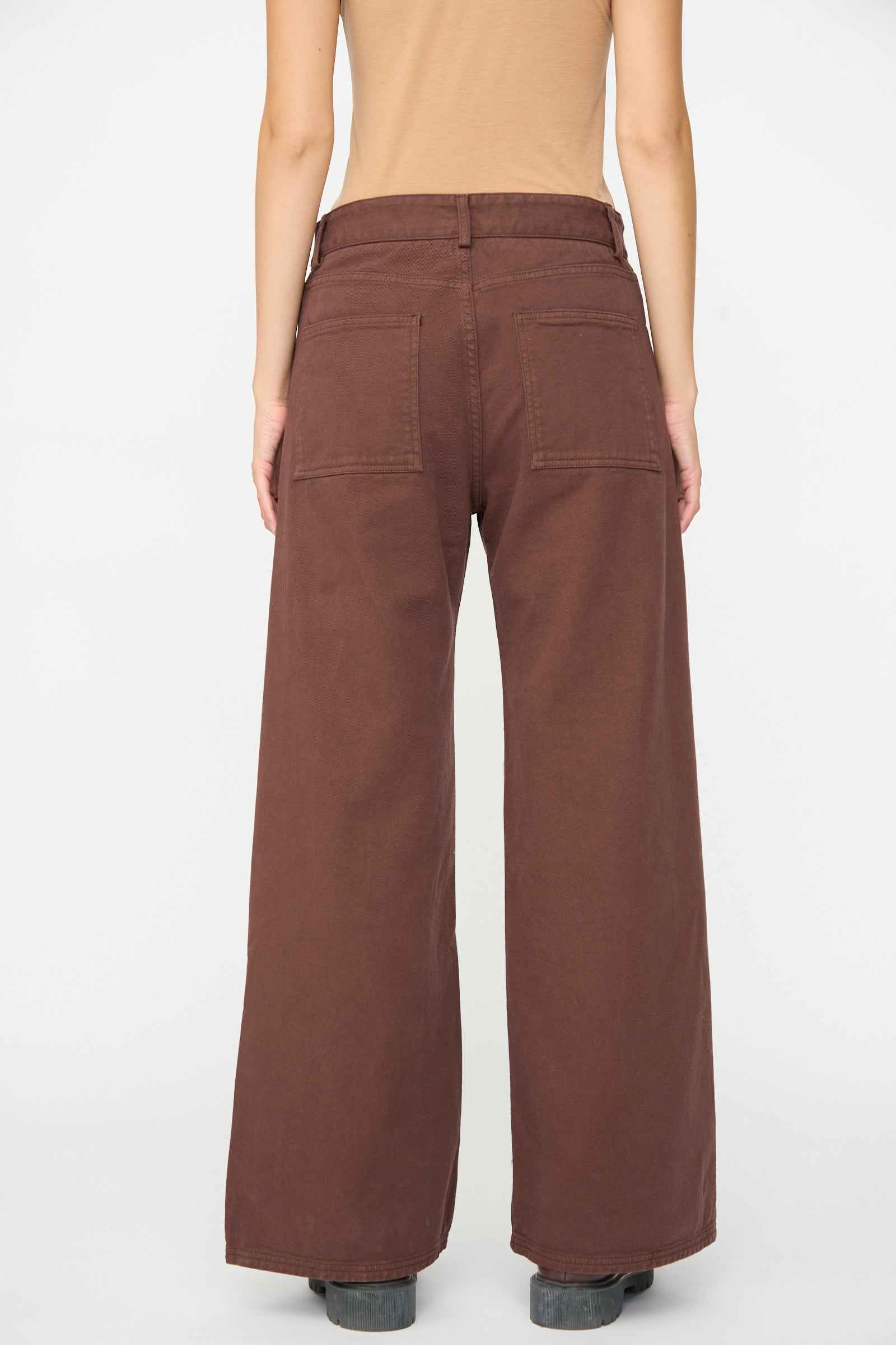 Dressed in the Tajo Pant Wide in Cata Brown by Baserange—crafted from gabardine cotton in Portugal—the individual stands with their back to the camera, paired with a beige top against a plain white background.