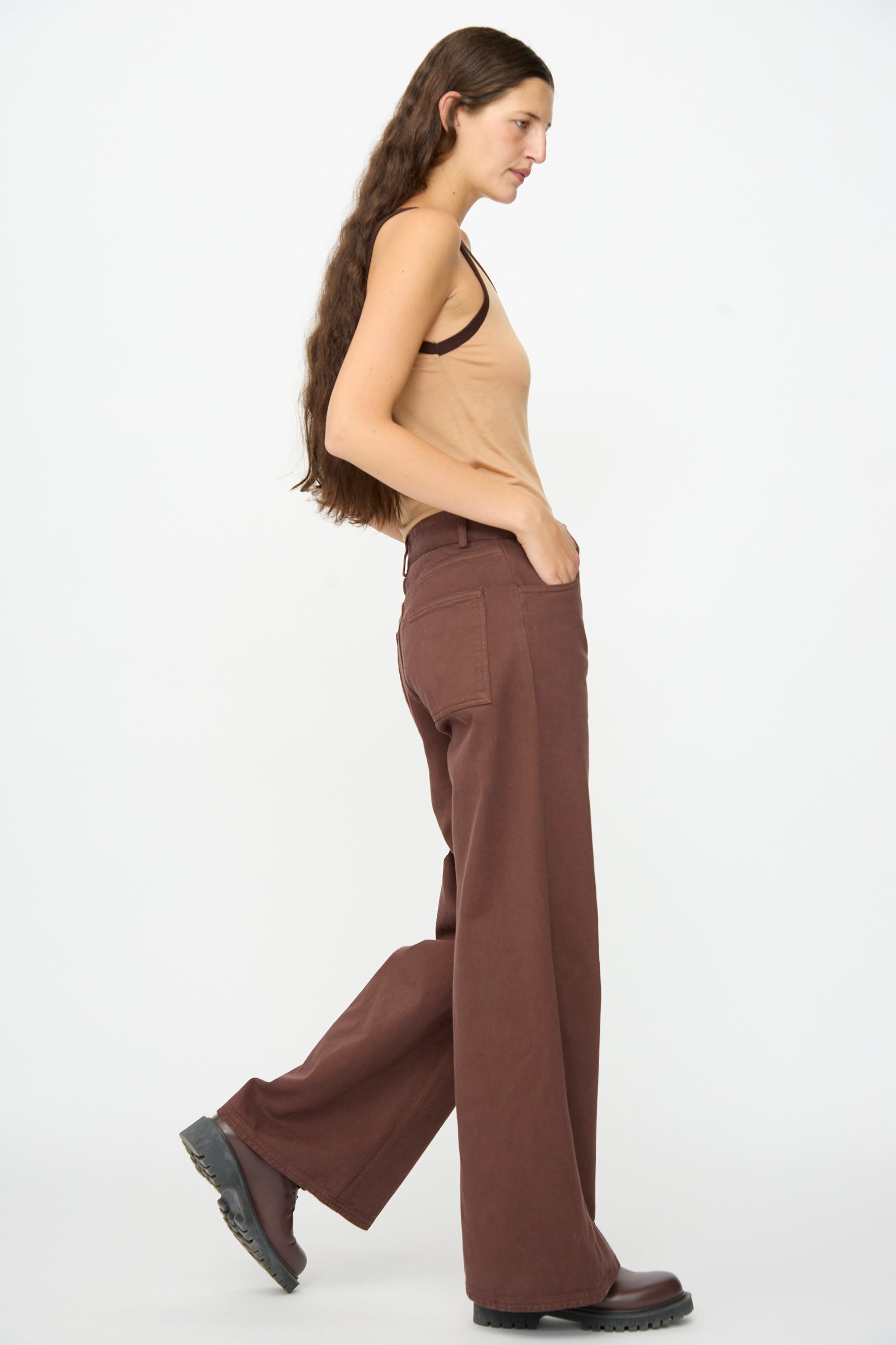 A woman with long brown hair wears a tan tank top and the Baserange Tajo Pant Wide in Cata Brown, along with dark shoes, walking sideways against a plain white background.