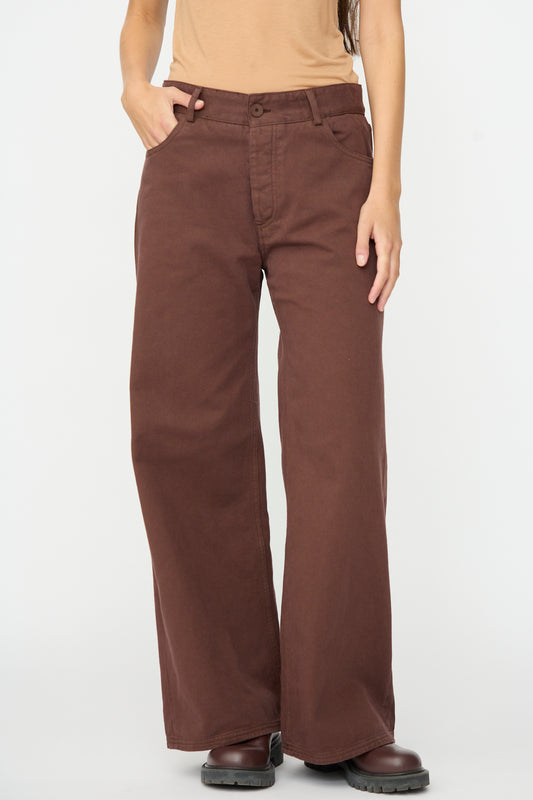 A person wearing the Baserange Tajo Pant Wide in Cata Brown, paired with a tan short-sleeve top and dark brown shoes, stands with one hand in their pocket against a plain white background.