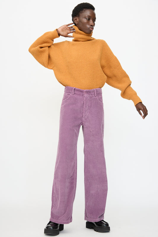 A person wearing a mustard yellow turtleneck, Baserange Tajo Wide Pants in Cue Purple, and black platform shoes stands against a plain white background.
