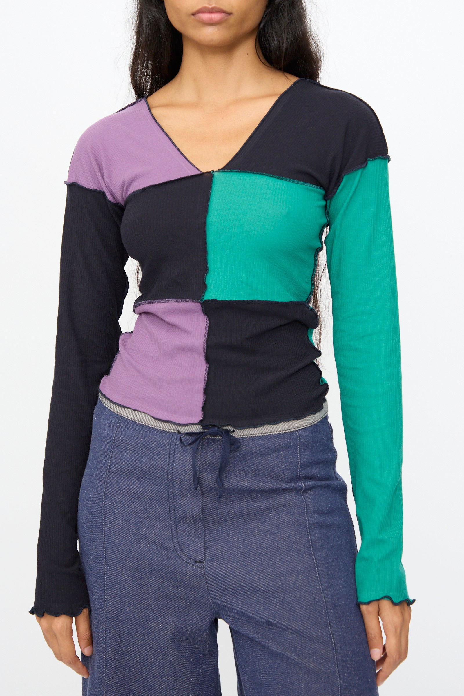 A woman wears the Baserange Teak Long Sleeve Tee in green, navy, and purple with high-waisted blue pants, standing against a plain white background.
