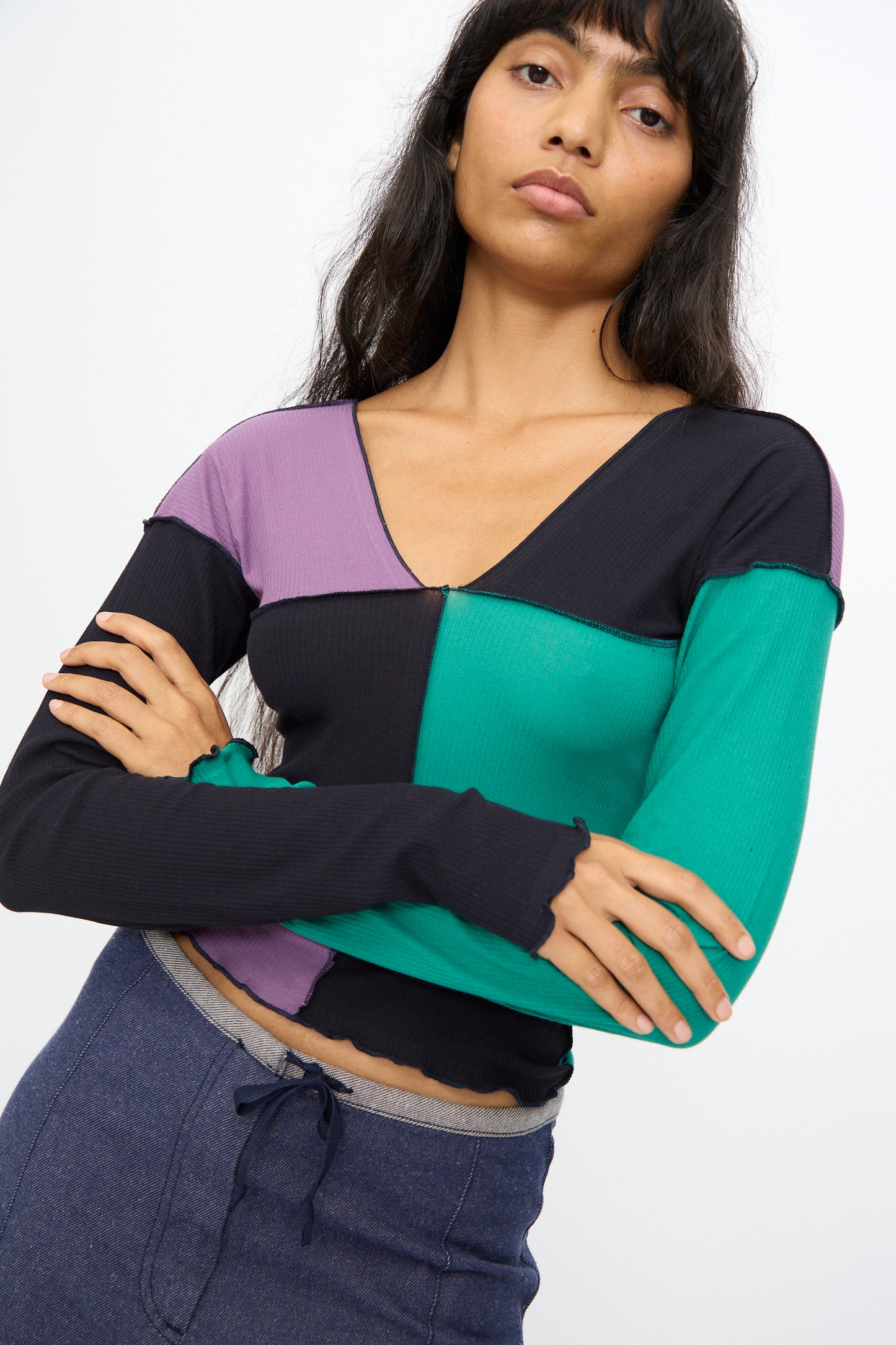 A woman with long dark hair wears the Baserange Teak Long Sleeve Tee in green, navy, and purple, paired with blue drawstring pants, standing with arms crossed.