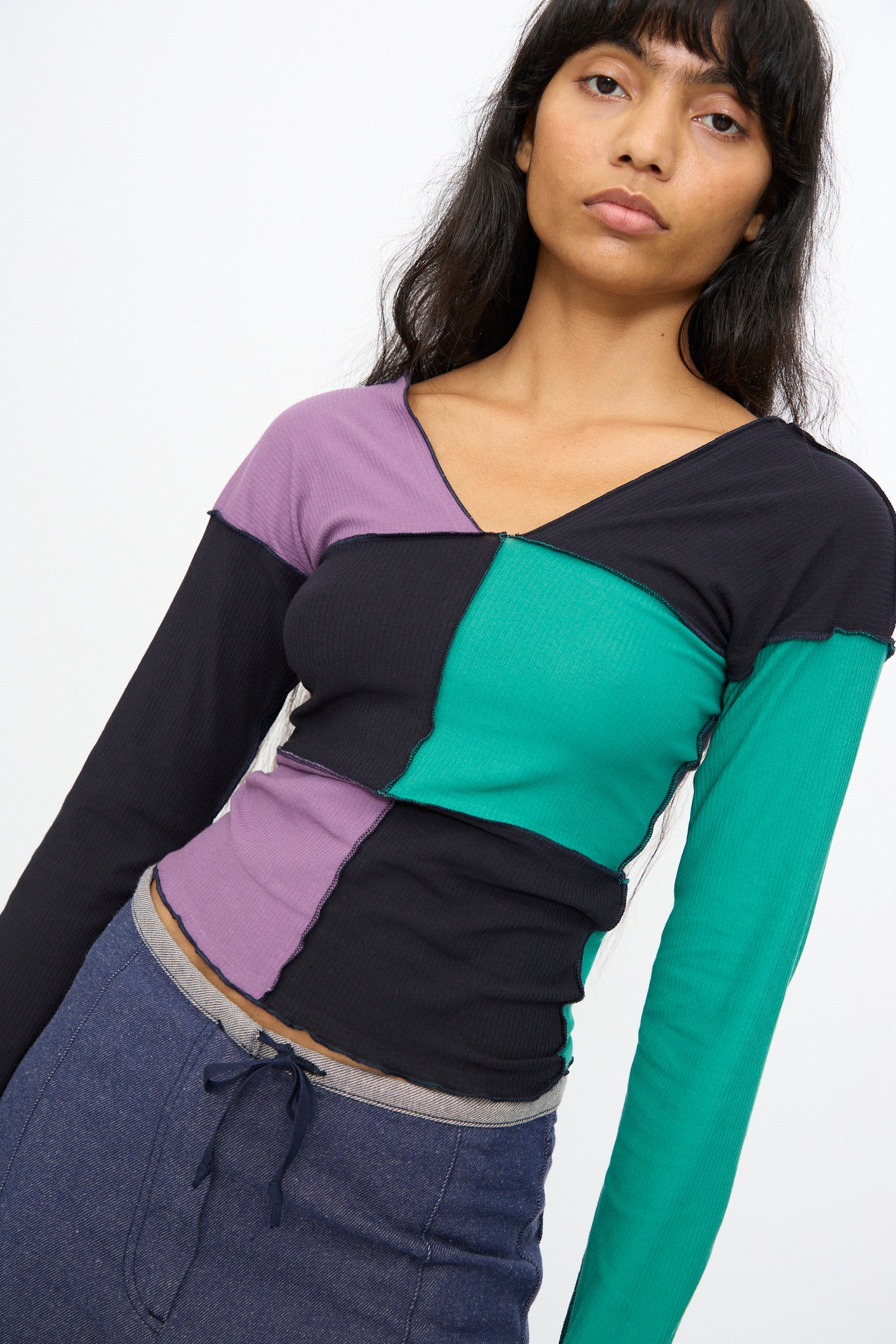 A woman wears the Baserange Teak Long Sleeve Tee in green, navy, and purple with visible seams, paired with blue drawstring pants, standing against a plain white background.