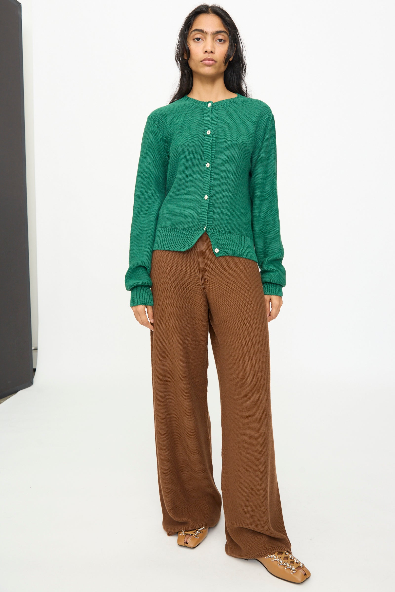 A person stands in a studio against a plain white background wearing the Baserange Tier Cardigan in Calla Green, styled with brown wide-leg pants and tan pointed shoes.