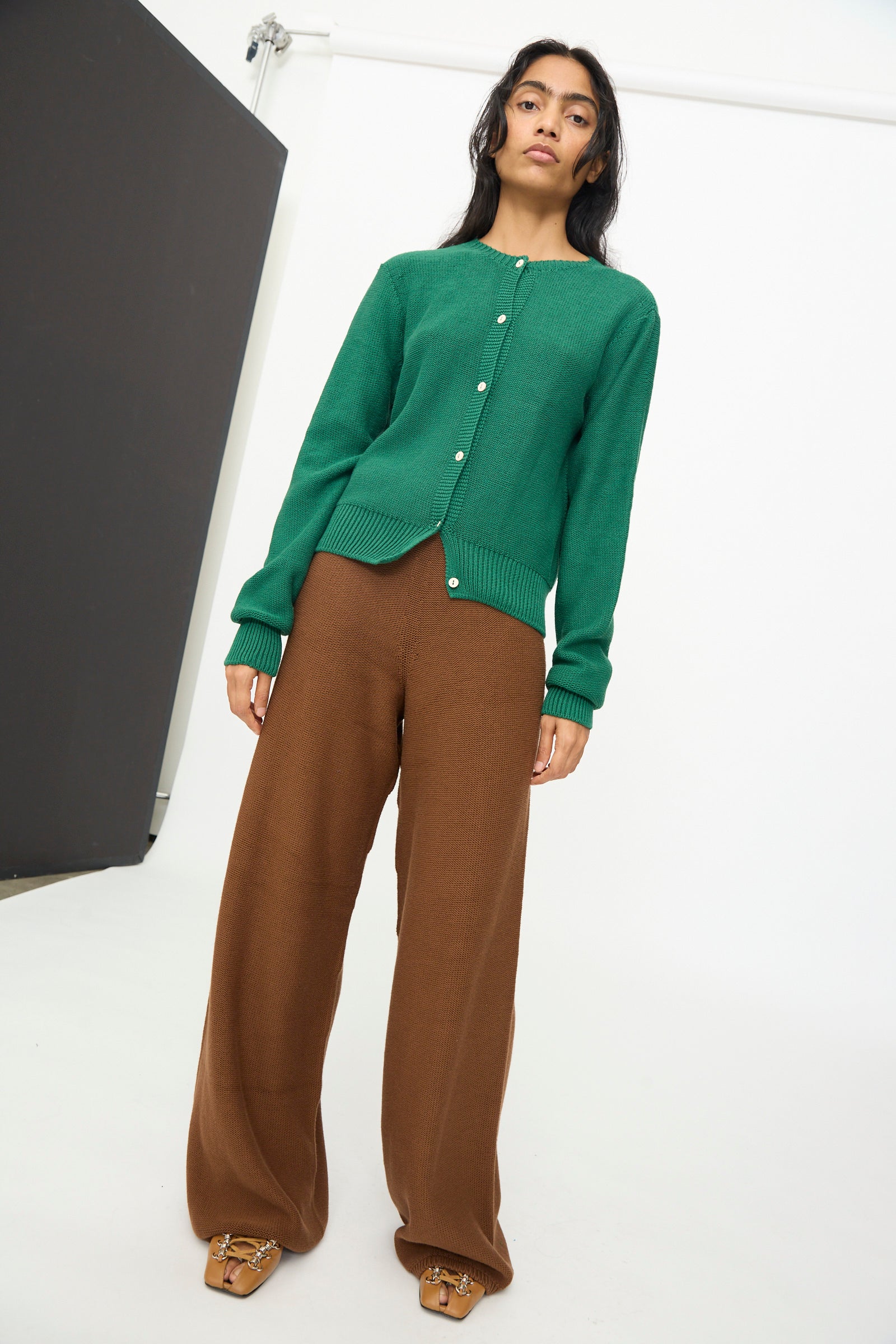 A woman stands in a studio with a neutral expression, wearing the Baserange Tier Cardigan in Calla Green, styled with wide brown pants and tan shoes.