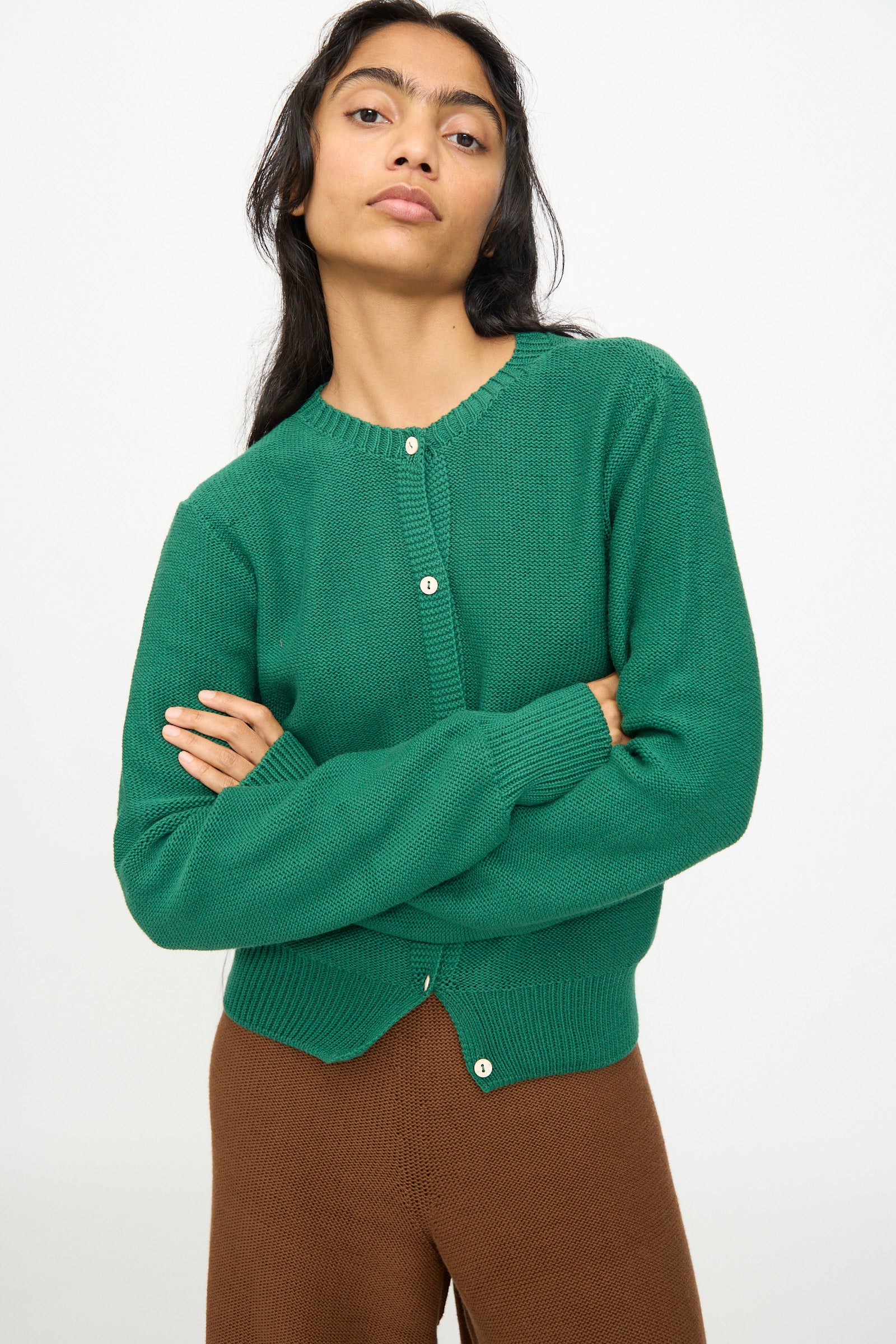 A person stands with arms crossed, wearing the Baserange Tier Cardigan in Calla Green and brown pants against a plain white background.