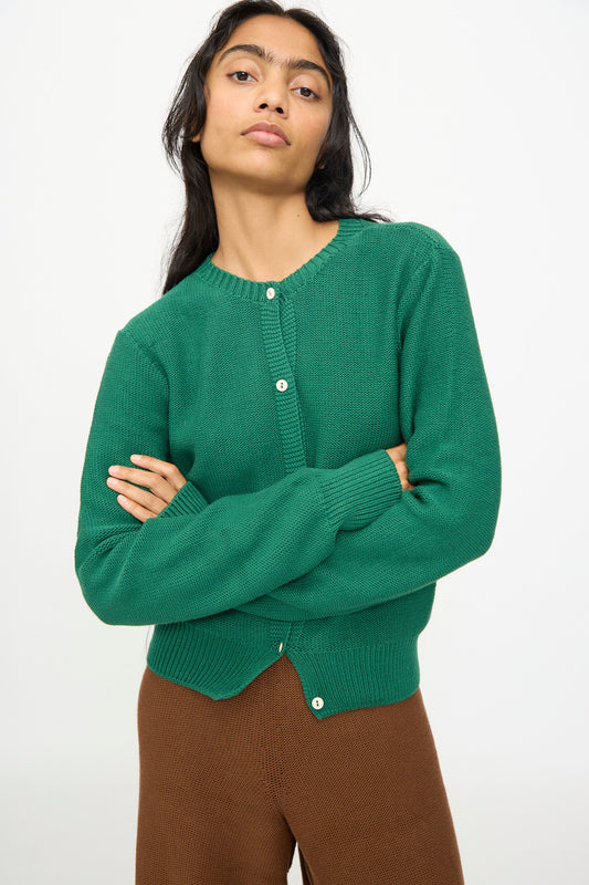 A person stands with arms crossed, wearing the Baserange Tier Cardigan in Calla Green and brown pants against a plain white background.