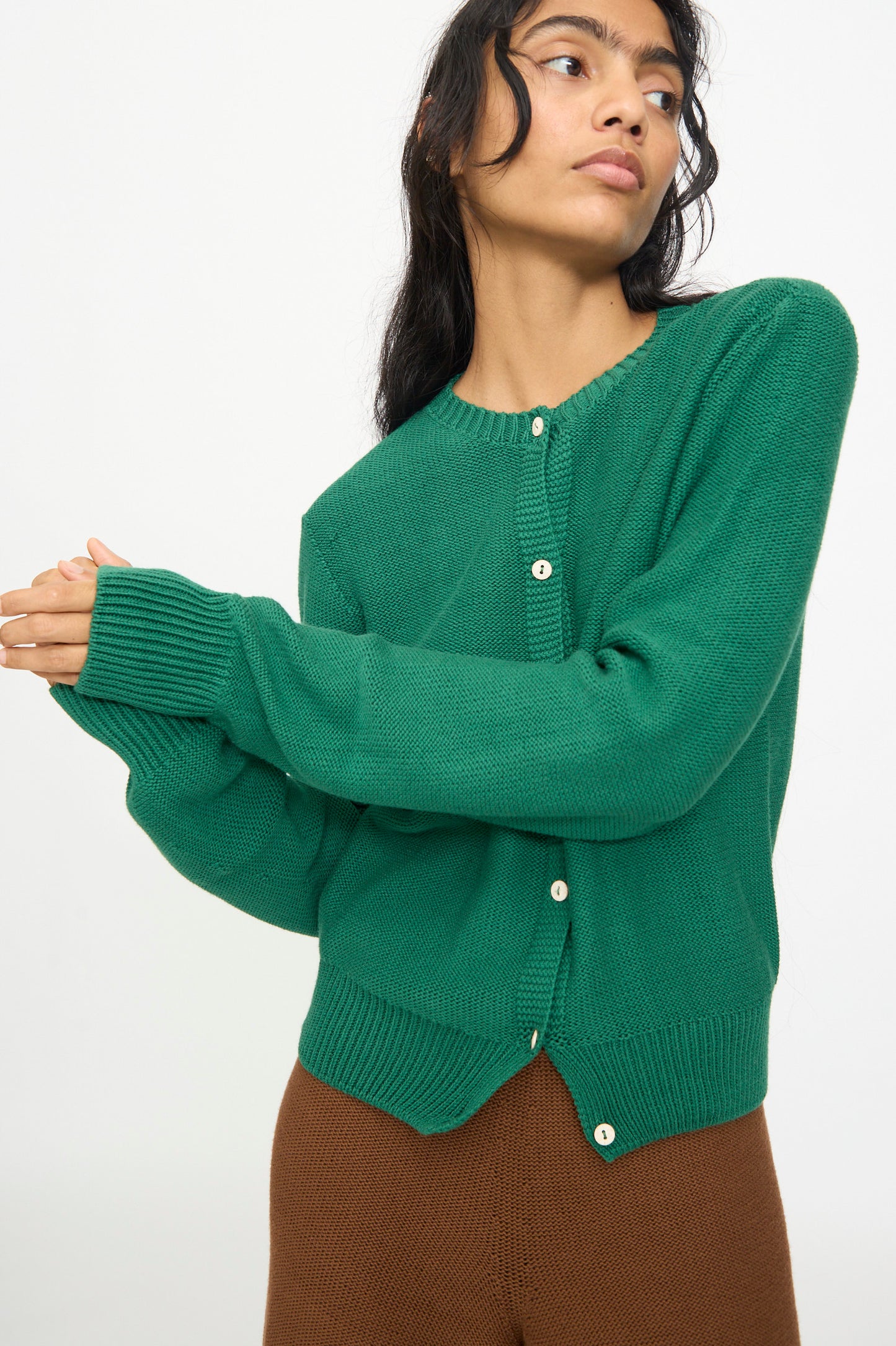 A woman wearing the Baserange Tier Cardigan in Calla Green—an organic cotton cropped cardigan—and brown pants, stands against a plain white background with her arms slightly extended.