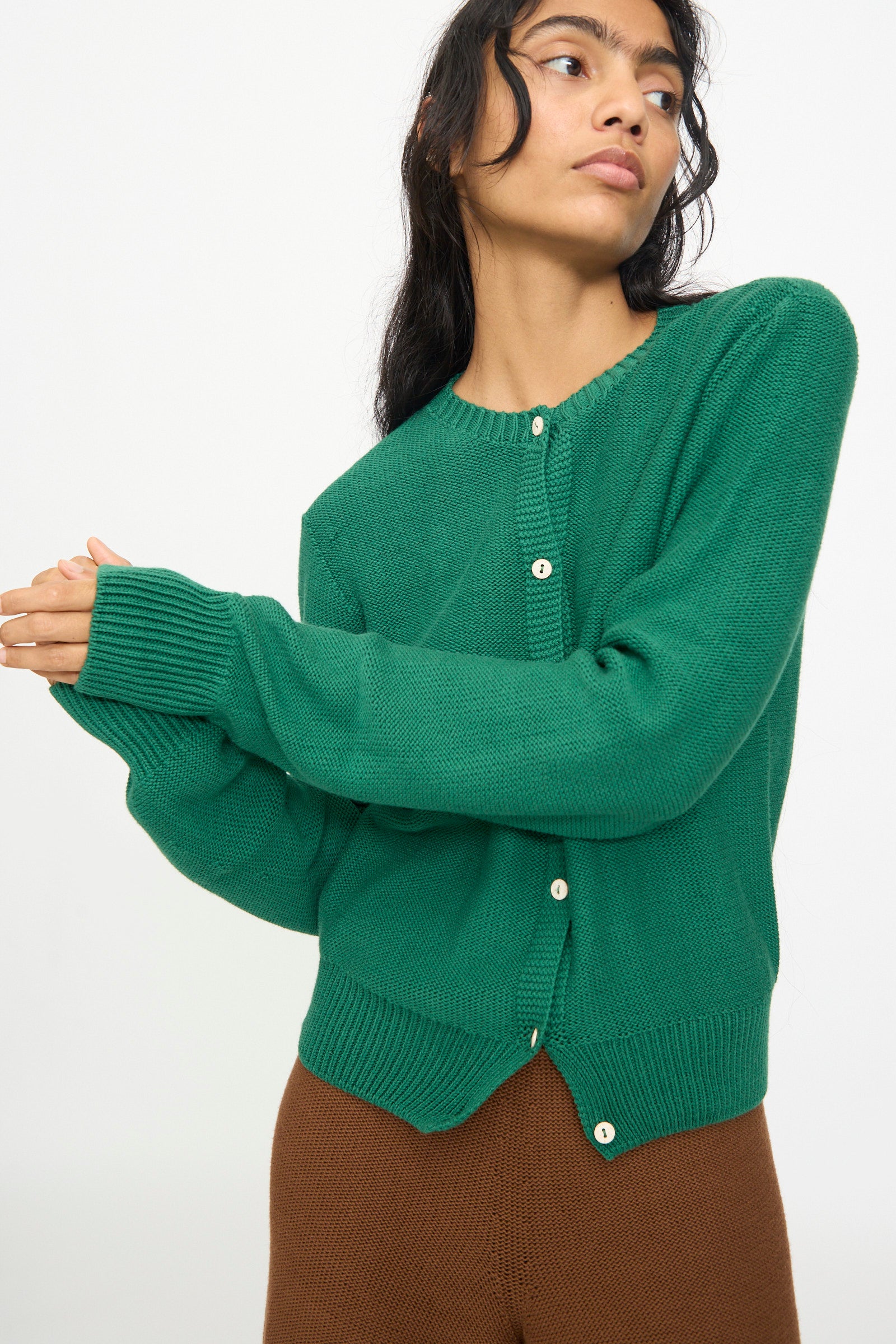 A woman wearing the Baserange Tier Cardigan in Calla Green—an organic cotton cropped cardigan—and brown pants, stands against a plain white background with her arms slightly extended.