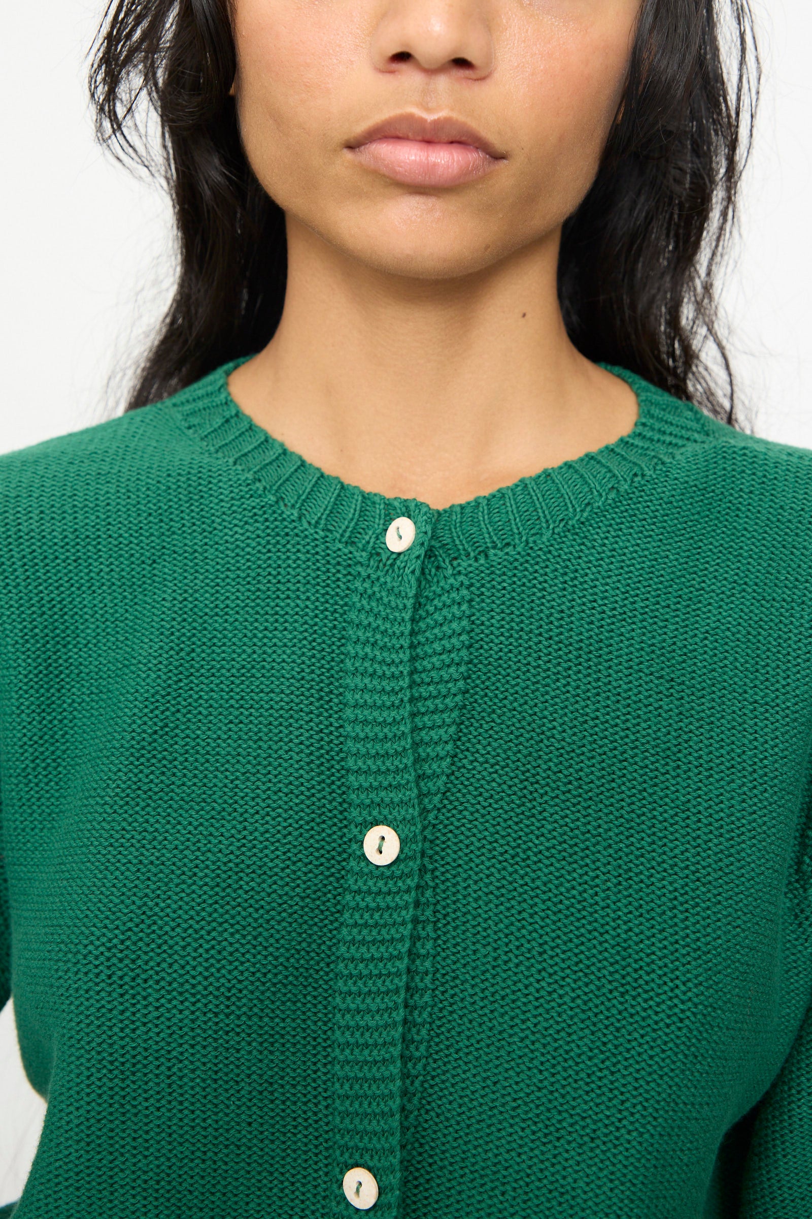A woman wearing the Baserange Tier Cardigan in Calla Green, an organic cotton textured cropped cardigan with cream-colored buttons, photographed from shoulders to mouth against a plain background.
