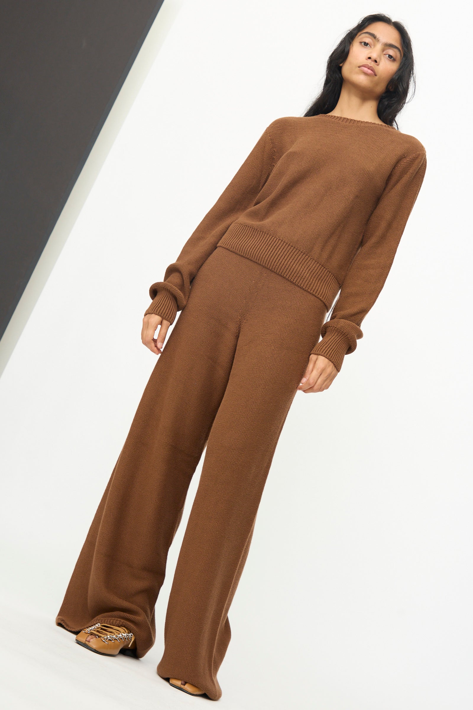 A person stands against a white wall, wearing Baserange's Tier Pants in Nocciola Brown—relaxed fit wide-leg pants crafted from organic cotton—paired with a matching brown knit long-sleeve sweater and brown shoes.