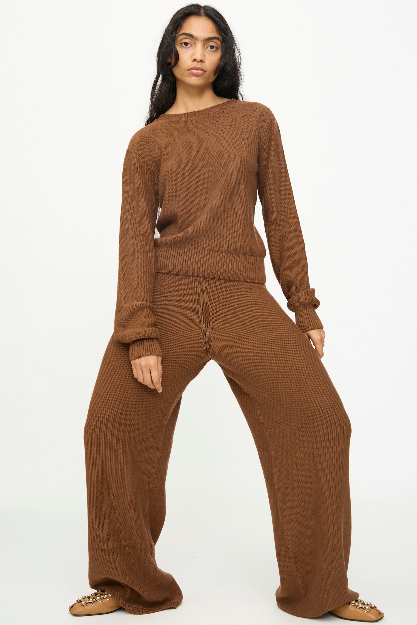 A person stands facing forward wearing the Baserange Tier Pants in Nocciola Brown, which are wide-leg, relaxed fit pants made from organic cotton, paired with a loose-fitting brown sweater and brown shoes against a plain light background.