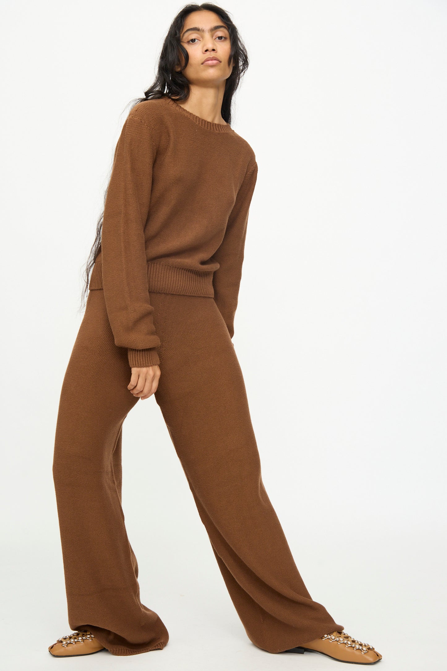 A person wearing the Baserange Tier Pants in Nocciola Brown, styled with a matching brown knit sweater, stands against a plain white background.