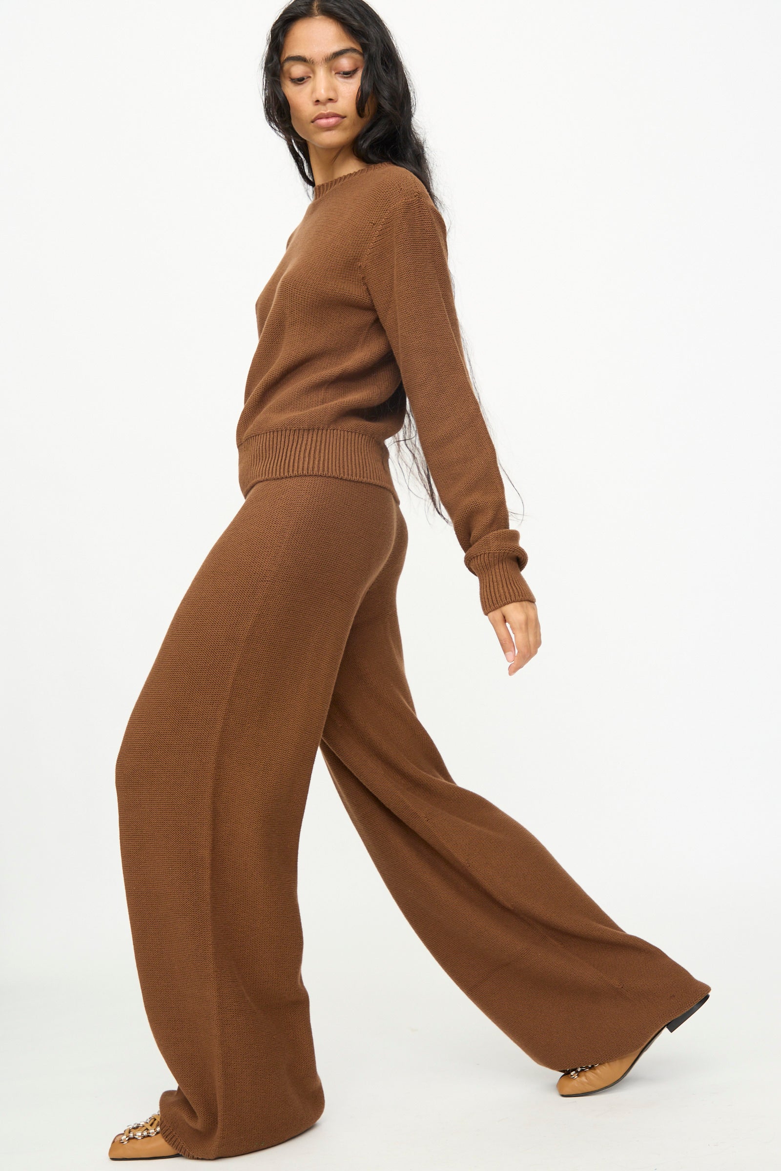 A woman wearing Baserange Tier Pants in Nocciola Brown, crafted from soft organic cotton with a relaxed wide-leg fit, walks against a plain white background.