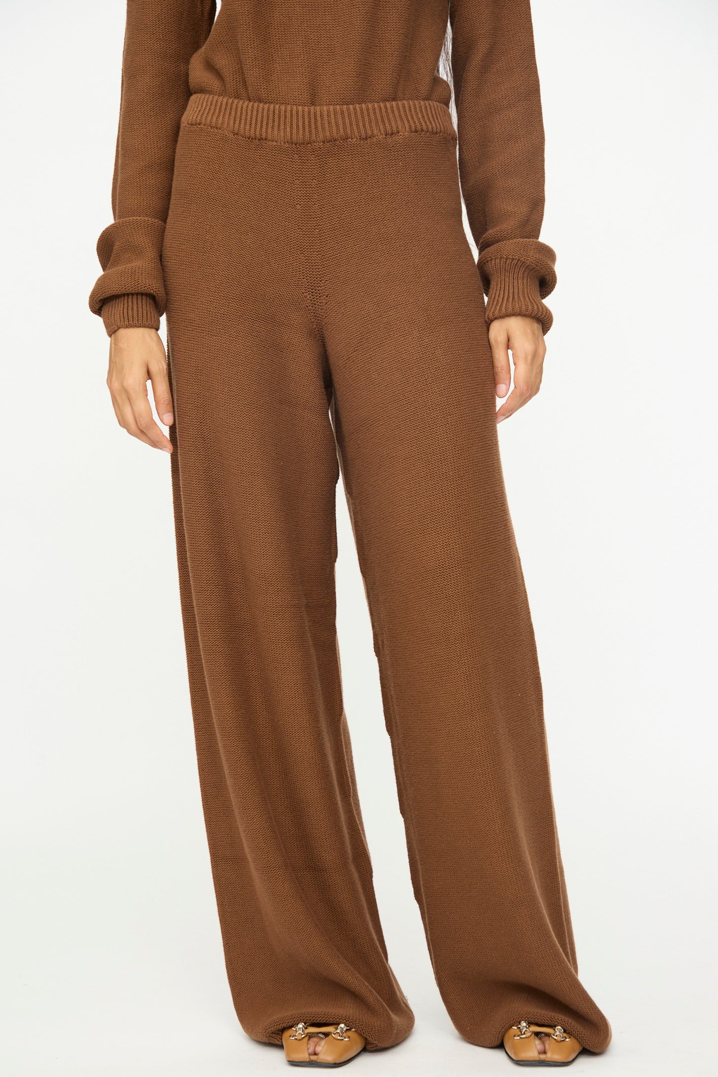 A person wearing Baserange's Tier Pants in Nocciola Brown, featuring an organic cotton wide-leg design with a relaxed fit, paired with a brown knit sweater and tan sandals, stands against a plain white background.