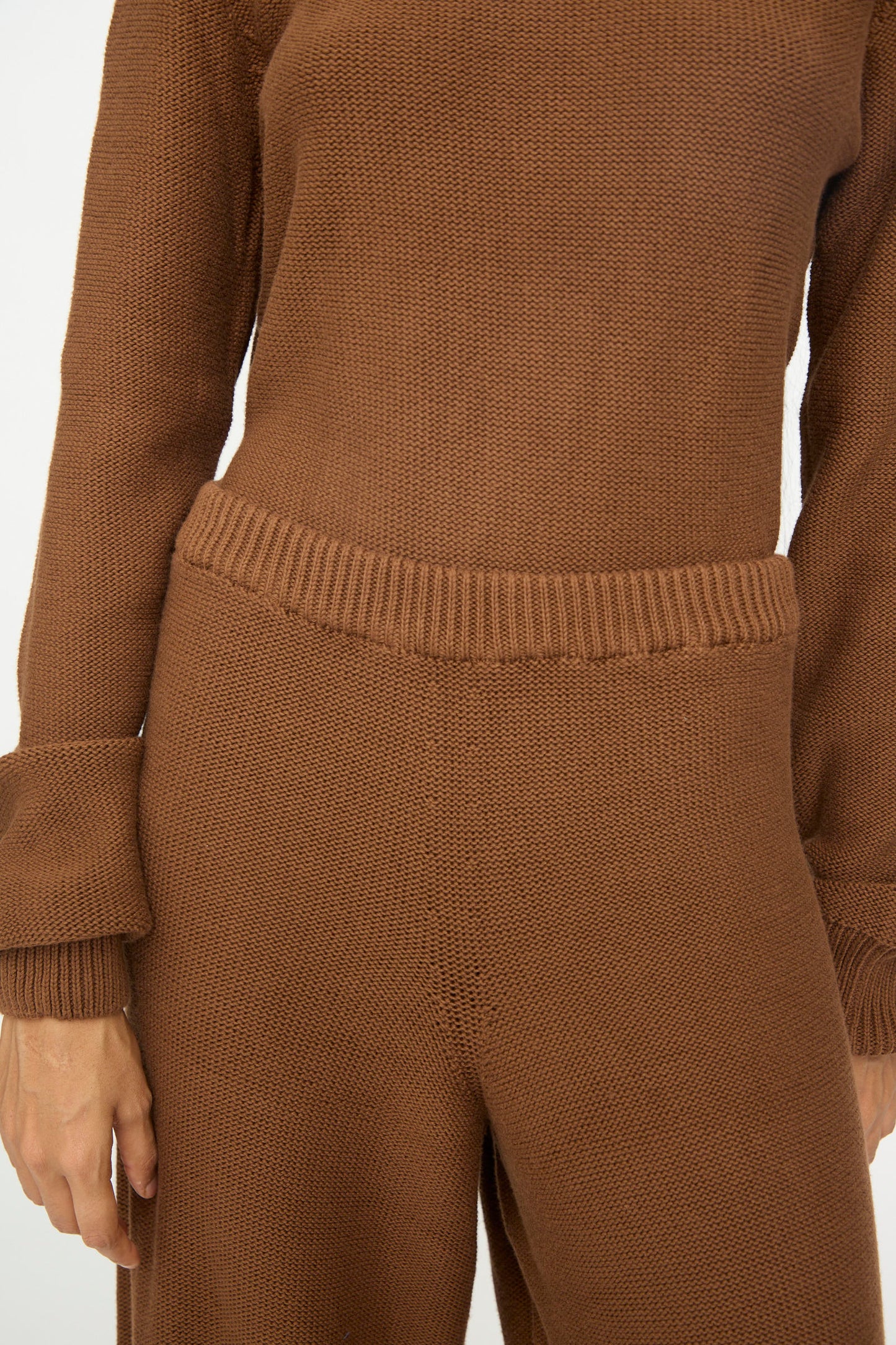 A person wearing the Baserange Tier Pants in Nocciola Brown, shown from shoulders to knees with arms relaxed at their sides, paired with a matching knit sweater.