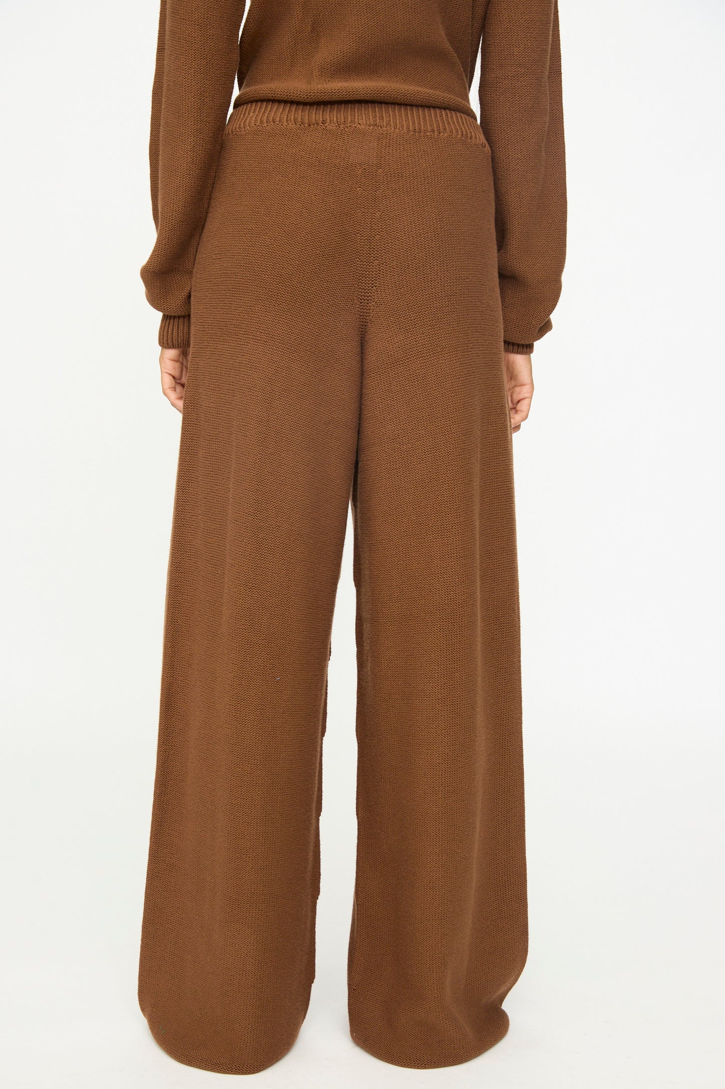A person is shown from the back against a plain white background, wearing Baserange's Tier Pants in Nocciola Brown—loose-fitting, high-waisted wide leg pants paired with a matching brown sweater. The relaxed fit and soft organic cotton fabric provide both comfort and effortless style.