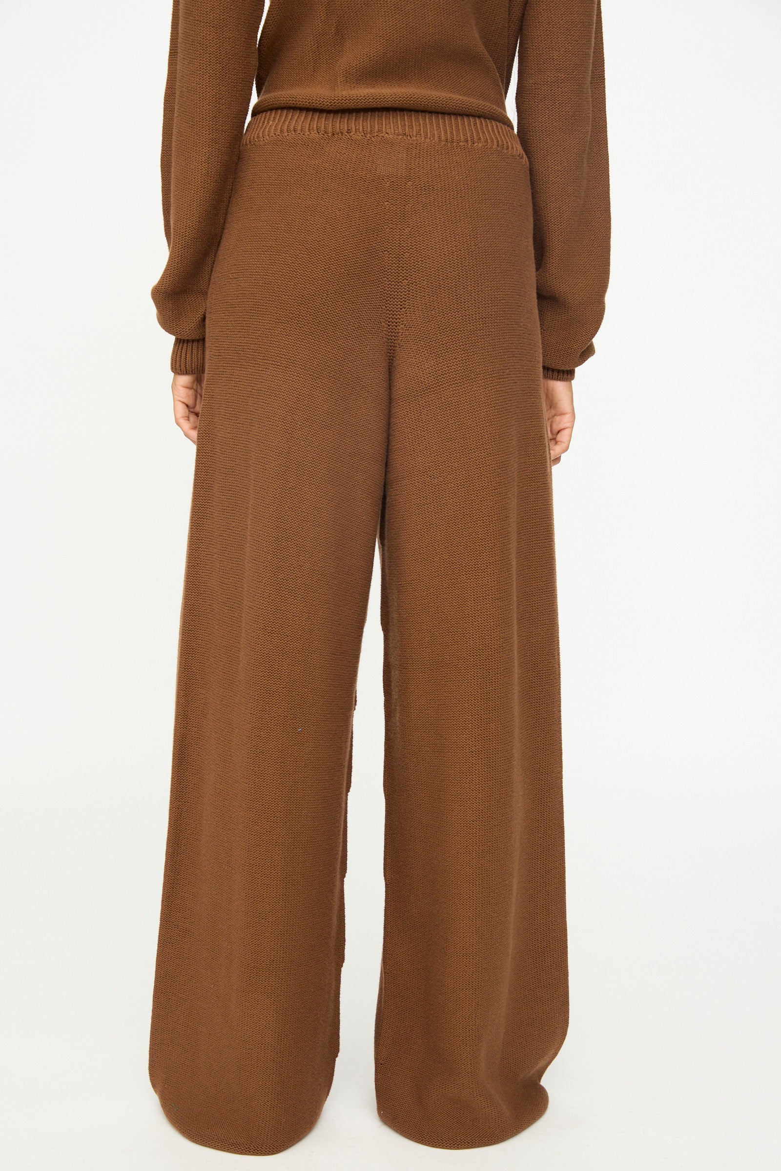A person is shown from the back against a plain white background, wearing Baserange's Tier Pants in Nocciola Brown—loose-fitting, high-waisted wide leg pants paired with a matching brown sweater. The relaxed fit and soft organic cotton fabric provide both comfort and effortless style.
