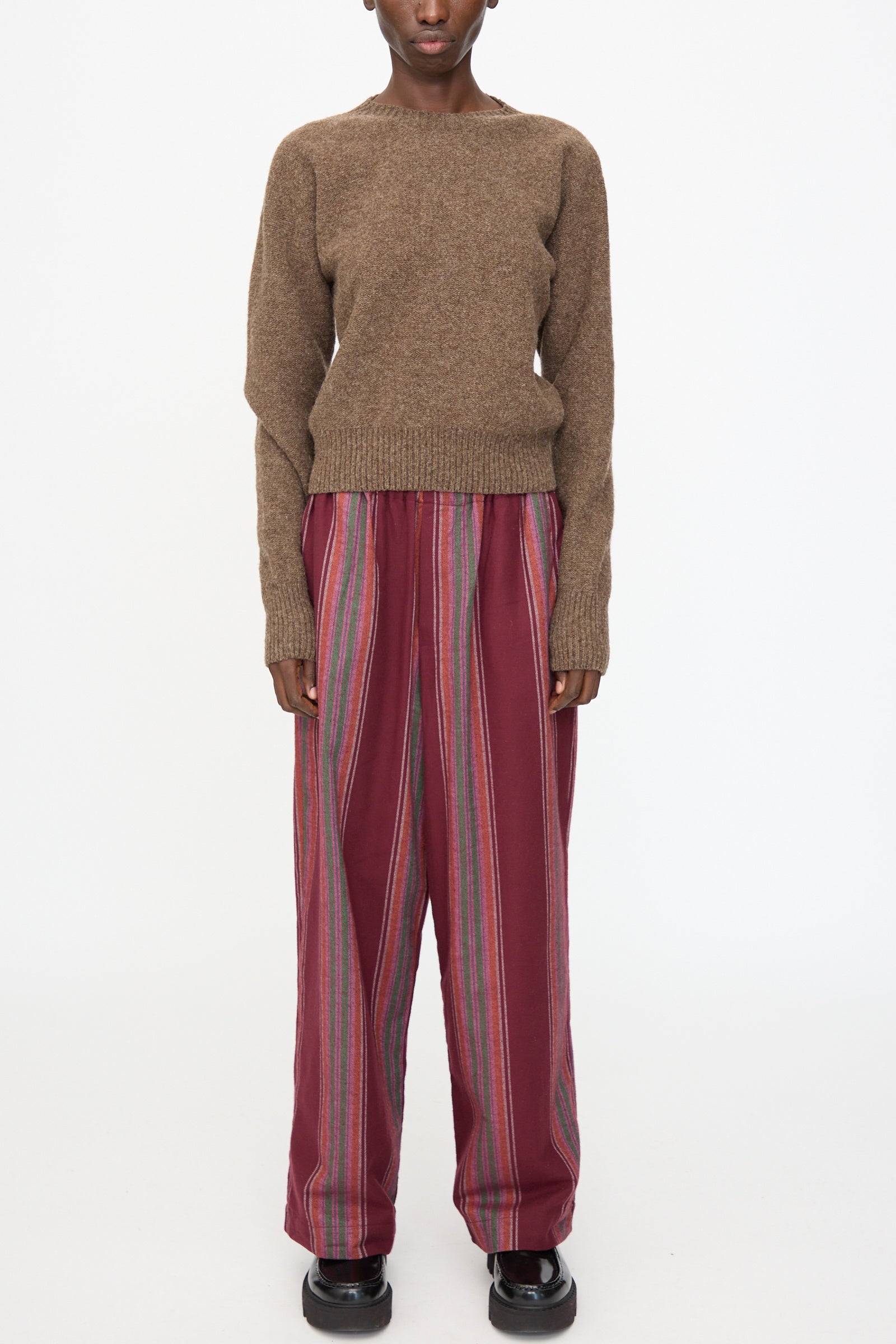 A model wears the Baserange Tier Sweater in Burel Brown, paired with loose maroon striped pants, standing against a plain white background.