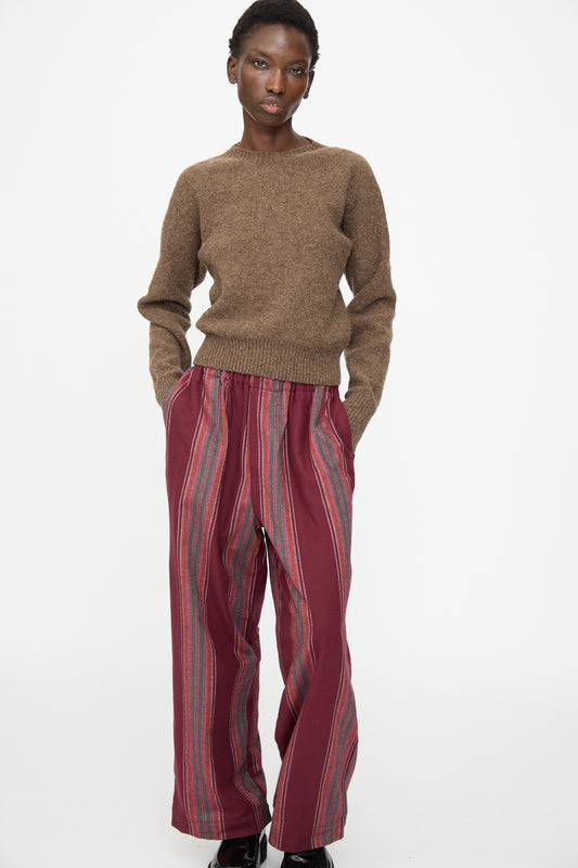 Person stands against a plain background, wearing a Baserange Tier Sweater in Burel Brown and loose maroon pants with vertical stripes.
