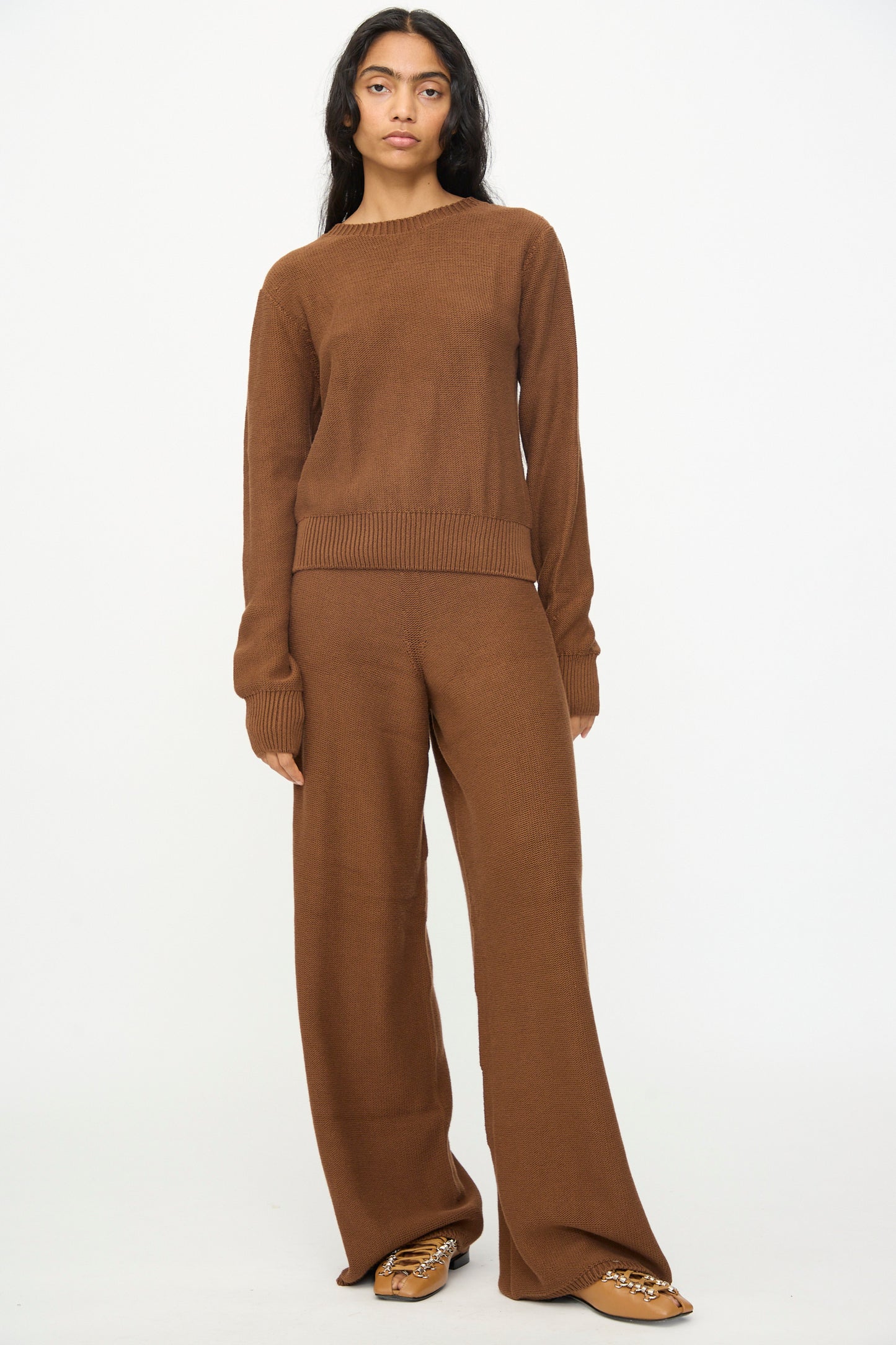 A person stands against a white background wearing the Baserange Tier Sweater in Nocciola Brown—an organic cotton pullover with a cropped fit—styled with wide-leg pants and tan embellished flat shoes.