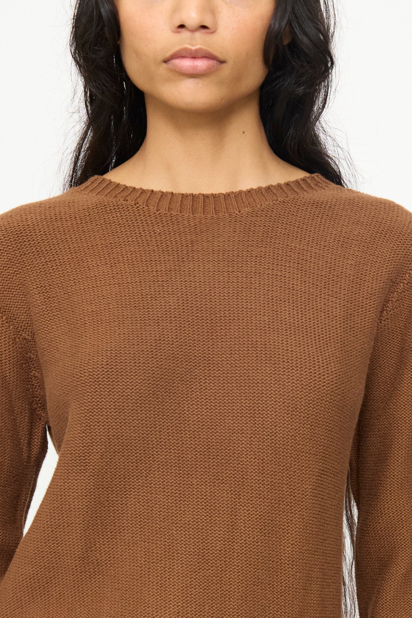 A person with long dark hair and a neutral expression is shown from shoulders to mid-torso wearing the Baserange Tier Sweater in Nocciola Brown.