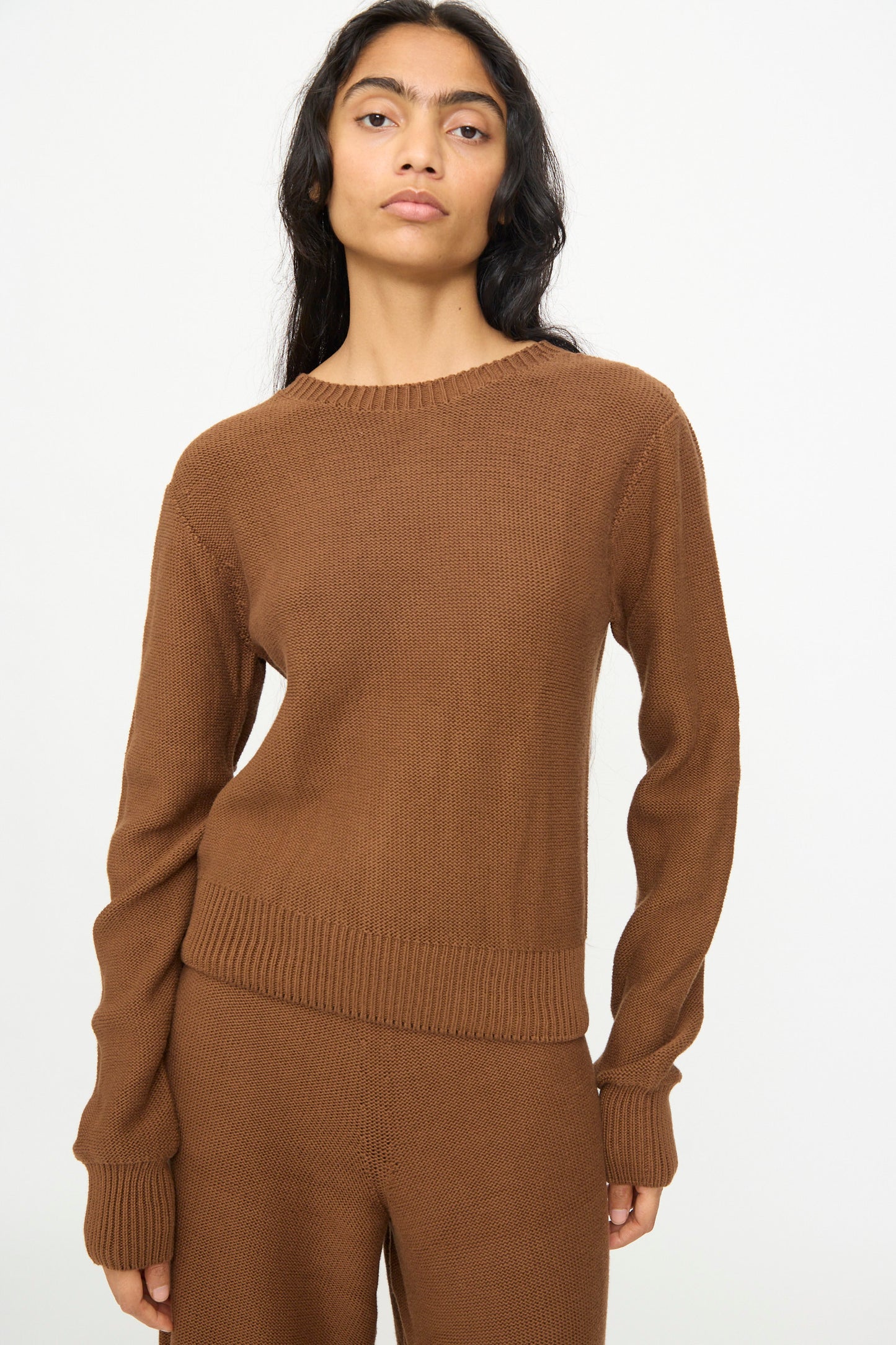 A person wearing the Baserange Tier Sweater in Nocciola Brown and matching brown knit pants stands against a plain white background, facing the camera with a neutral expression.