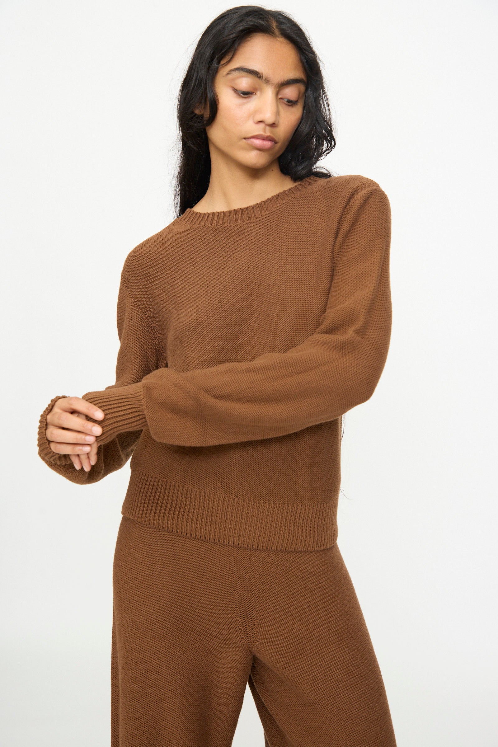 A person with long dark hair wears the Baserange Tier Sweater in Nocciola Brown—a cropped pullover—paired with matching pants, standing against a plain light background.