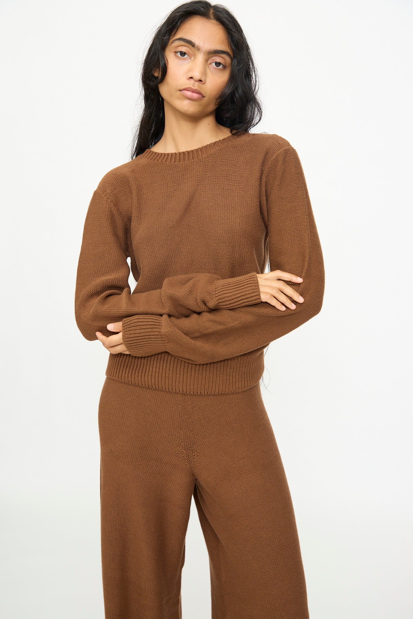 Person standing against a plain background, wearing the Baserange Tier Sweater in Nocciola Brown and matching pants, with arms loosely crossed and a neutral facial expression.