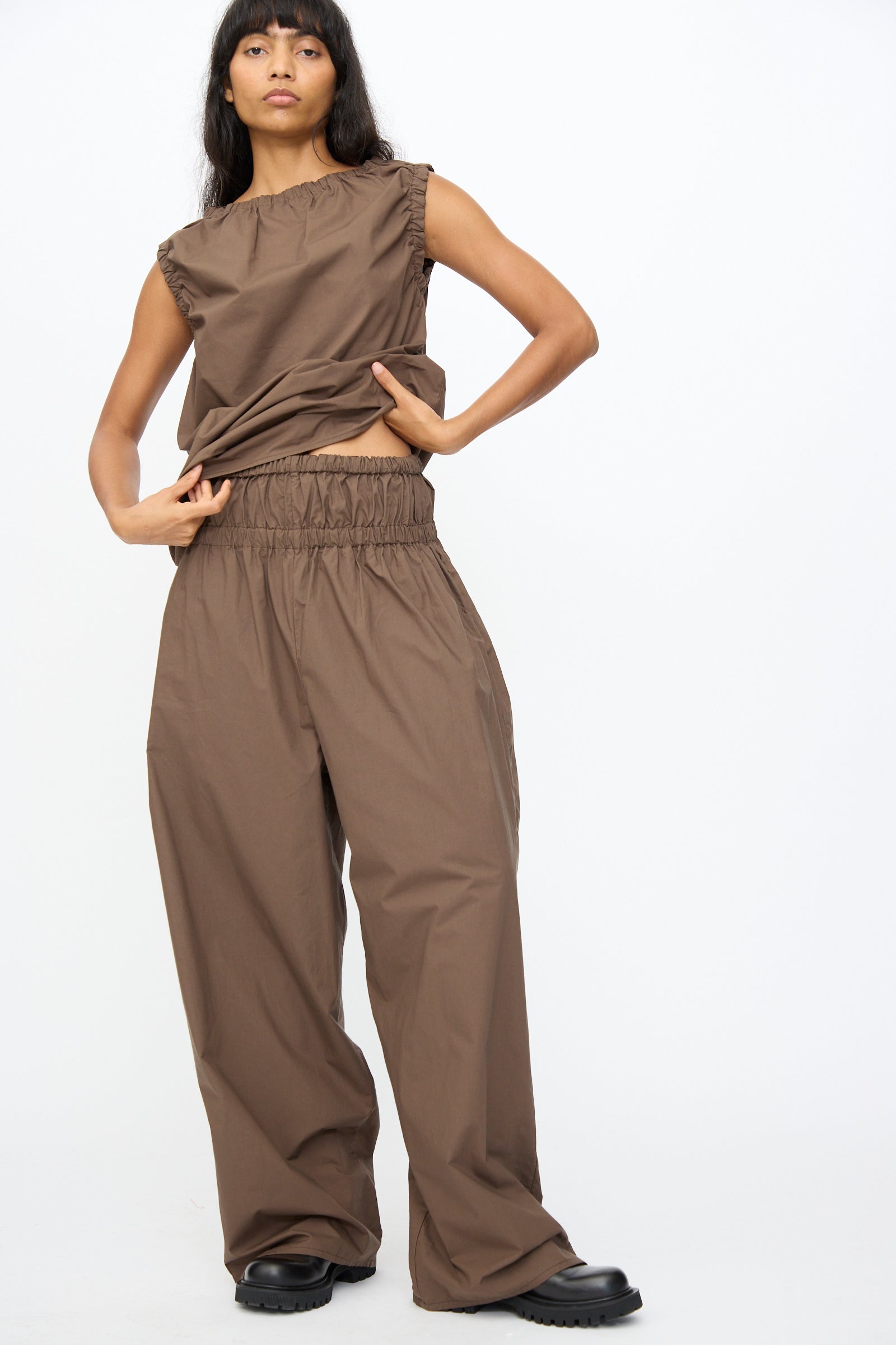 A person stands against a plain white background wearing Baserange’s Tin Pant in Mara Brown—loose, oversized organic cotton pants—paired with a matching brown sleeveless top and black shoes.