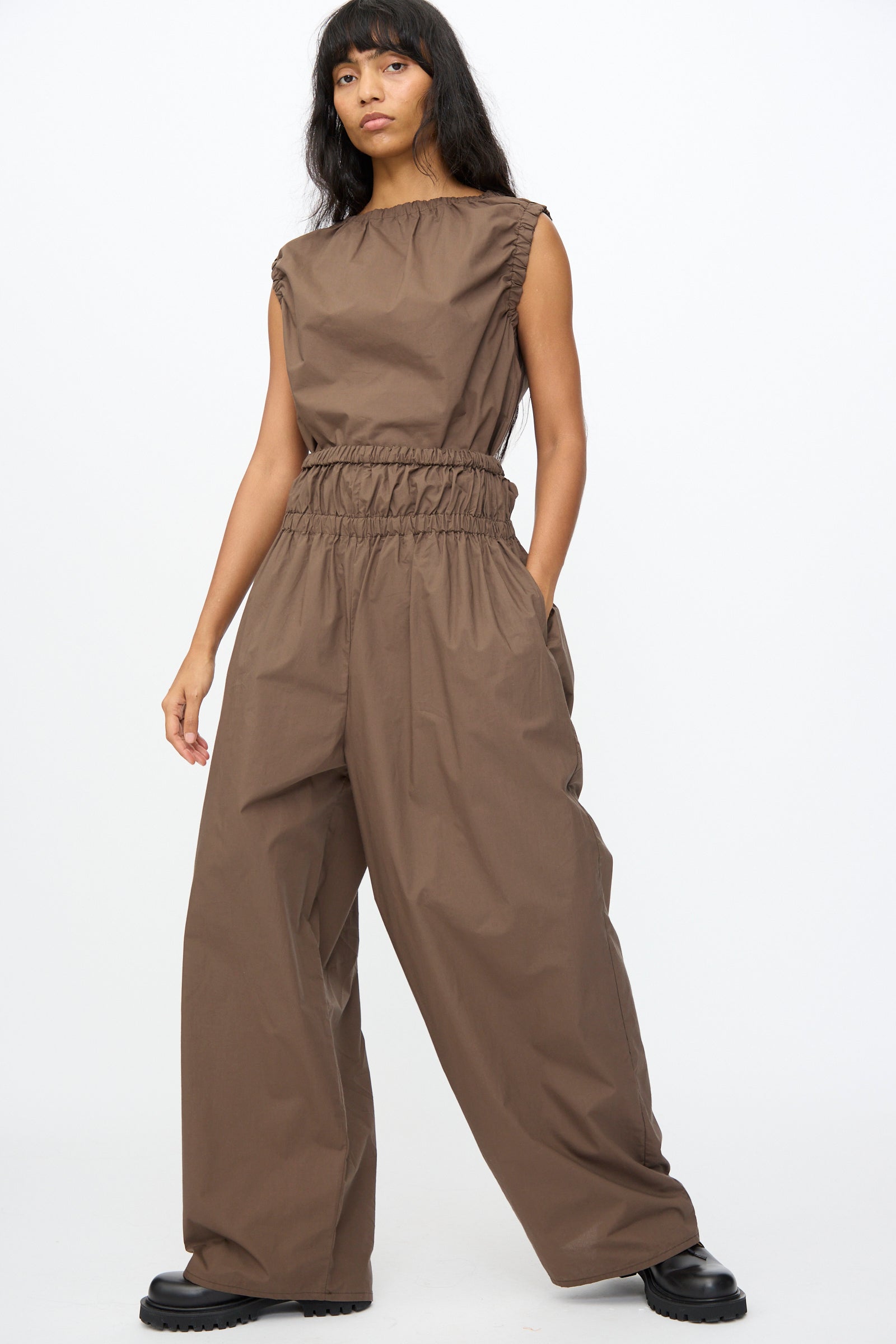 A person wears the Baserange Tin Pant in Mara Brown with oversized legs and black shoes, standing against a plain white background.