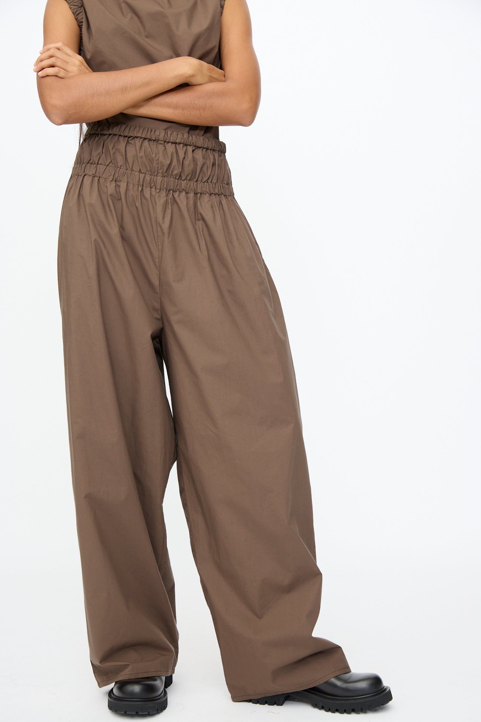 Person wearing Baserange Tin Pant in Mara Brown—oversized organic cotton pants with elastic waistband—paired with black shoes, arms crossed, upper face not visible, against a plain white background.