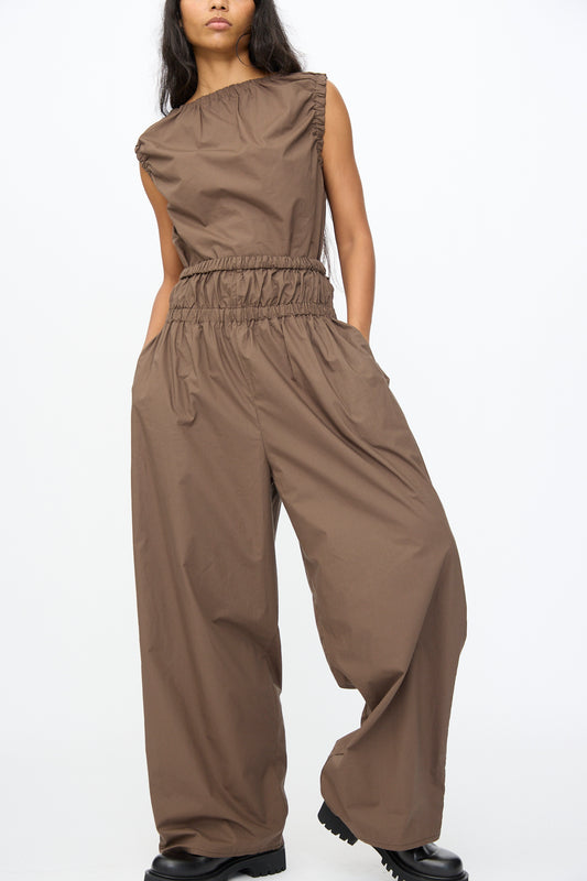 A person stands against a plain white background wearing Baserange’s Tin Pant in Mara Brown, an oversized cotton poplin jumpsuit with a gathered waistband and wide legs, styled with black chunky shoes.
