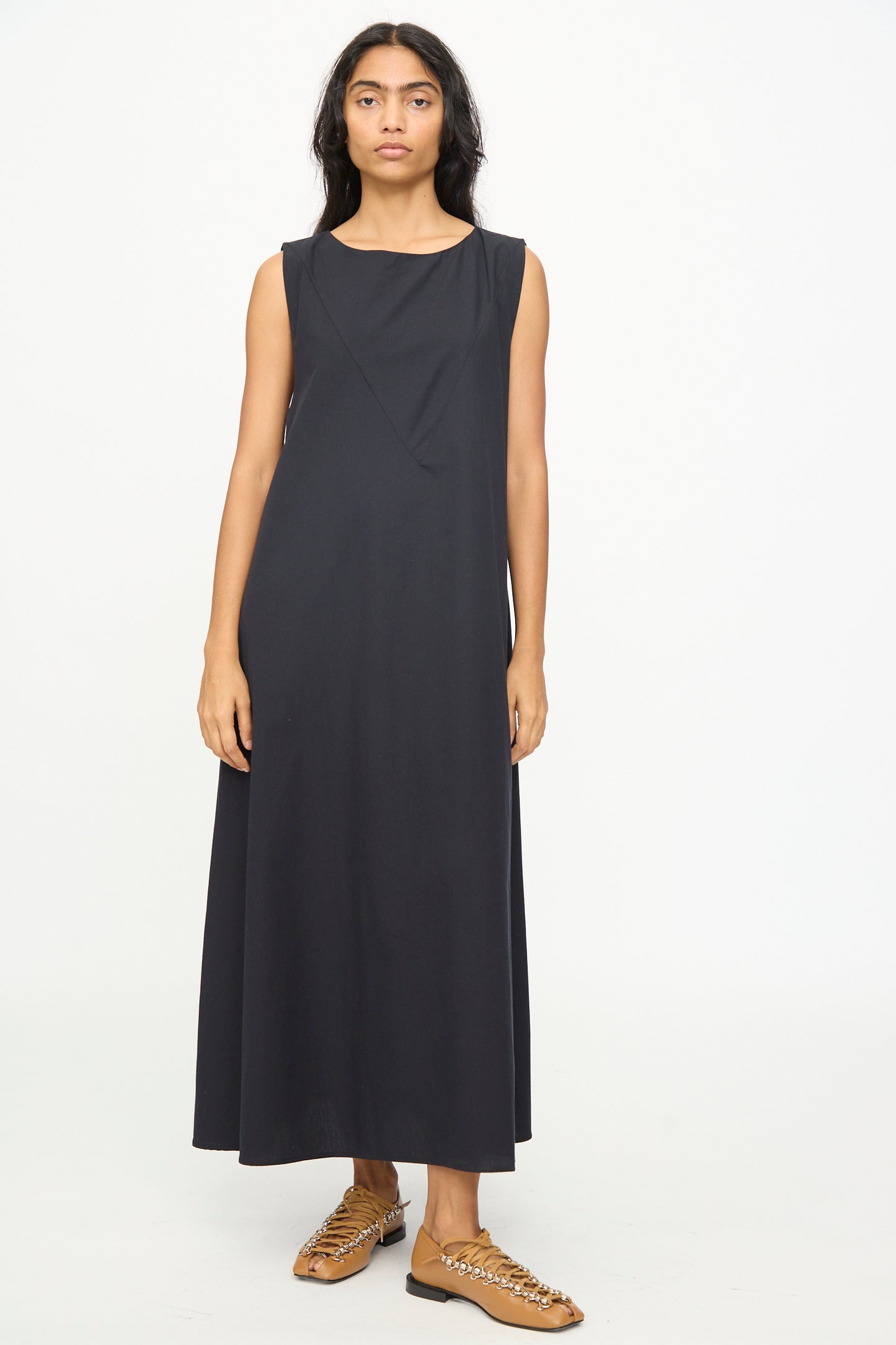 A person stands facing forward against a plain white background, wearing the Baserange Tres Dress in Black—a sleeveless, relaxed-fit dress—paired with brown woven shoes.