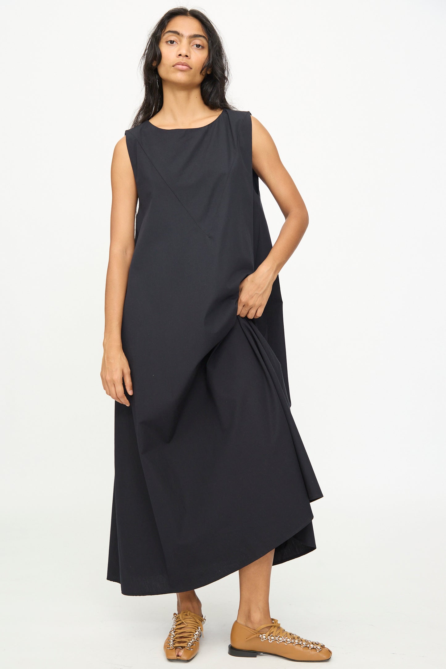 A person stands against a plain background wearing the Baserange Tres Dress in Black, styled with tan shoes featuring decorative embellishments.