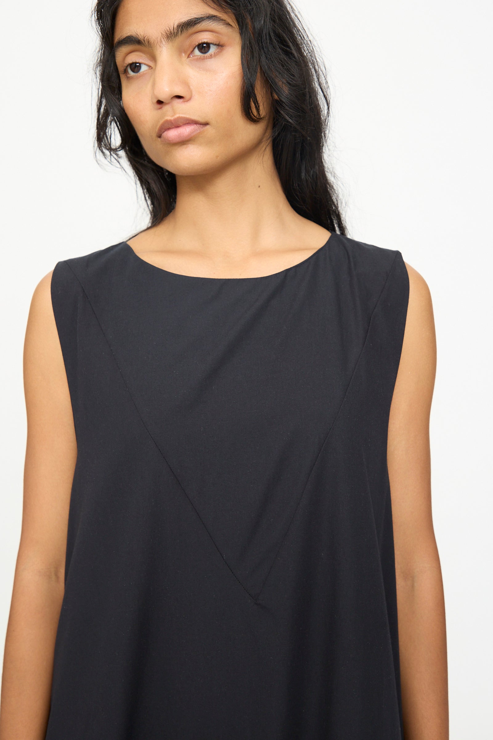 A woman with long dark hair wears the Baserange Tres Dress in Black, a sleeveless style featuring subtle seam detailing, standing against a plain light background.