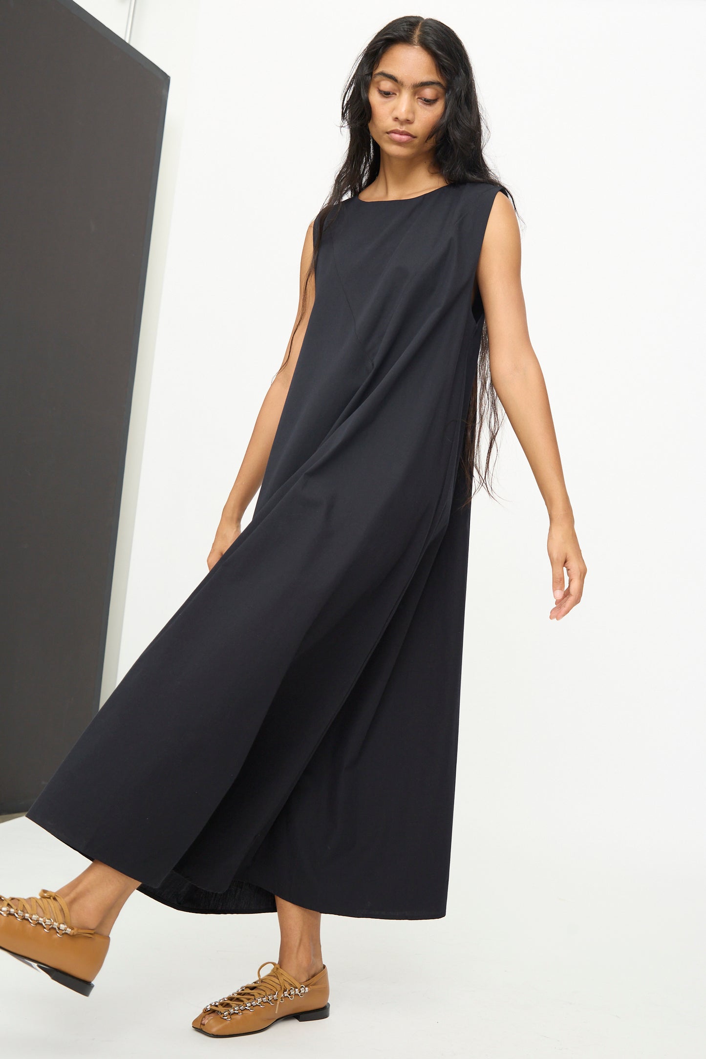 A woman stands against a plain, minimalist background wearing the Baserange Tres Dress in Black—a relaxed-fit, sleeveless dress crafted from cotton poplin—paired with brown loafers.