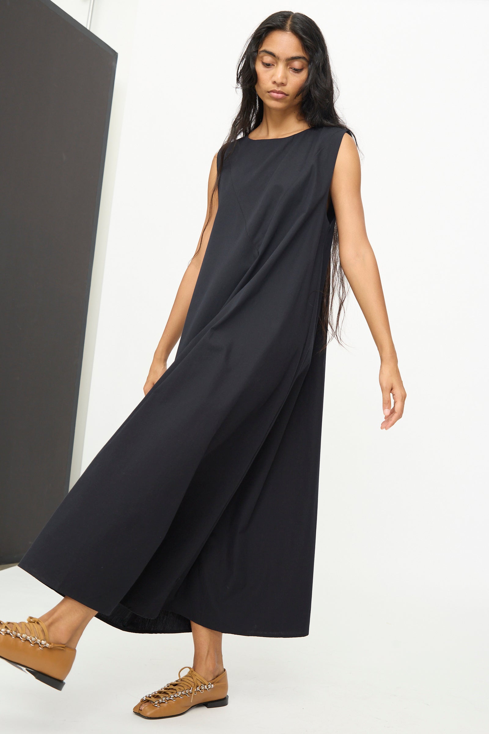A woman stands against a plain, minimalist background wearing the Baserange Tres Dress in Black—a relaxed-fit, sleeveless dress crafted from cotton poplin—paired with brown loafers.