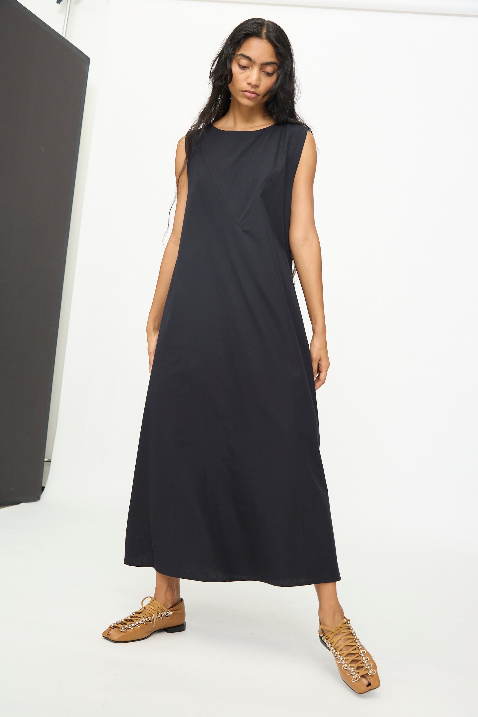 A woman stands in a studio against a plain white background, wearing the Baserange Tres Dress in Black—a sleeveless, relaxed-fit dress—paired with tan woven shoes.