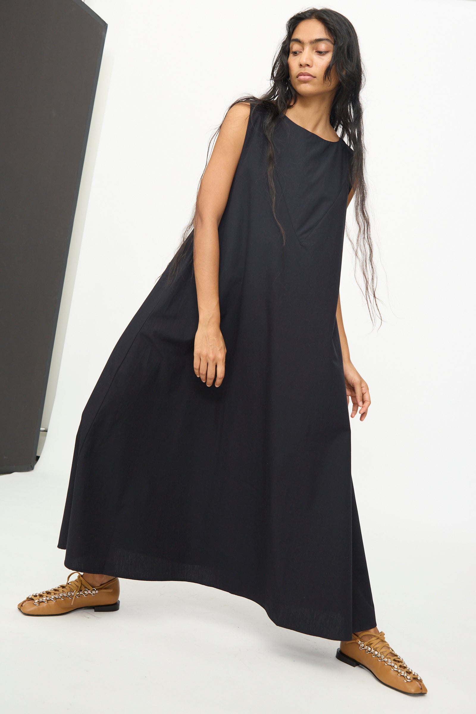 A person wearing the Baserange Tres Dress in Black—a long, sleeveless dress with a relaxed fit—and tan lace-up shoes stands against a plain white background.