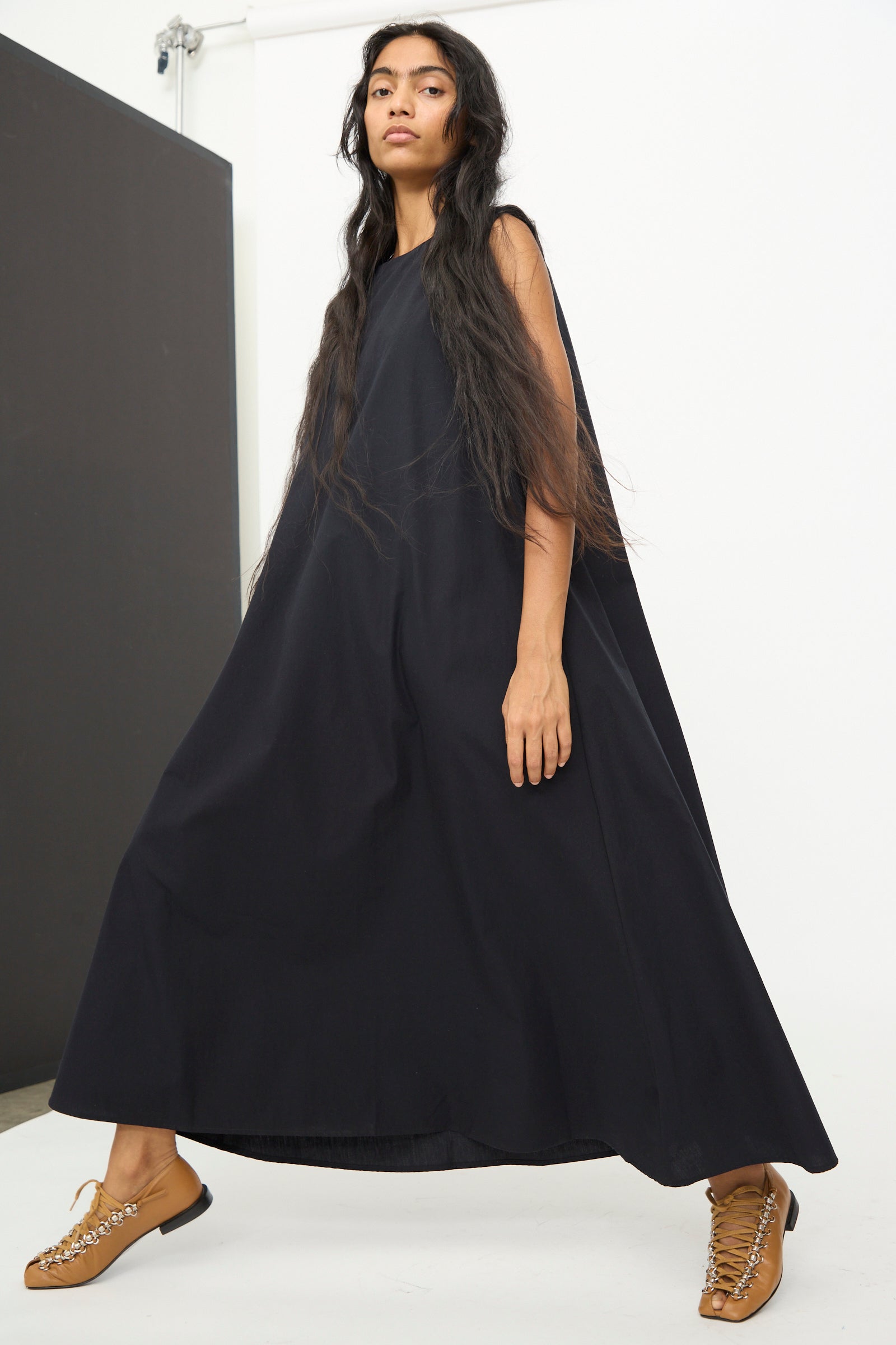 A woman wearing the Baserange Tres Dress in Black, a sleeveless garment with a relaxed fit, pairs it with tan lace-up shoes and stands against a white wall featuring a black panel in the background.