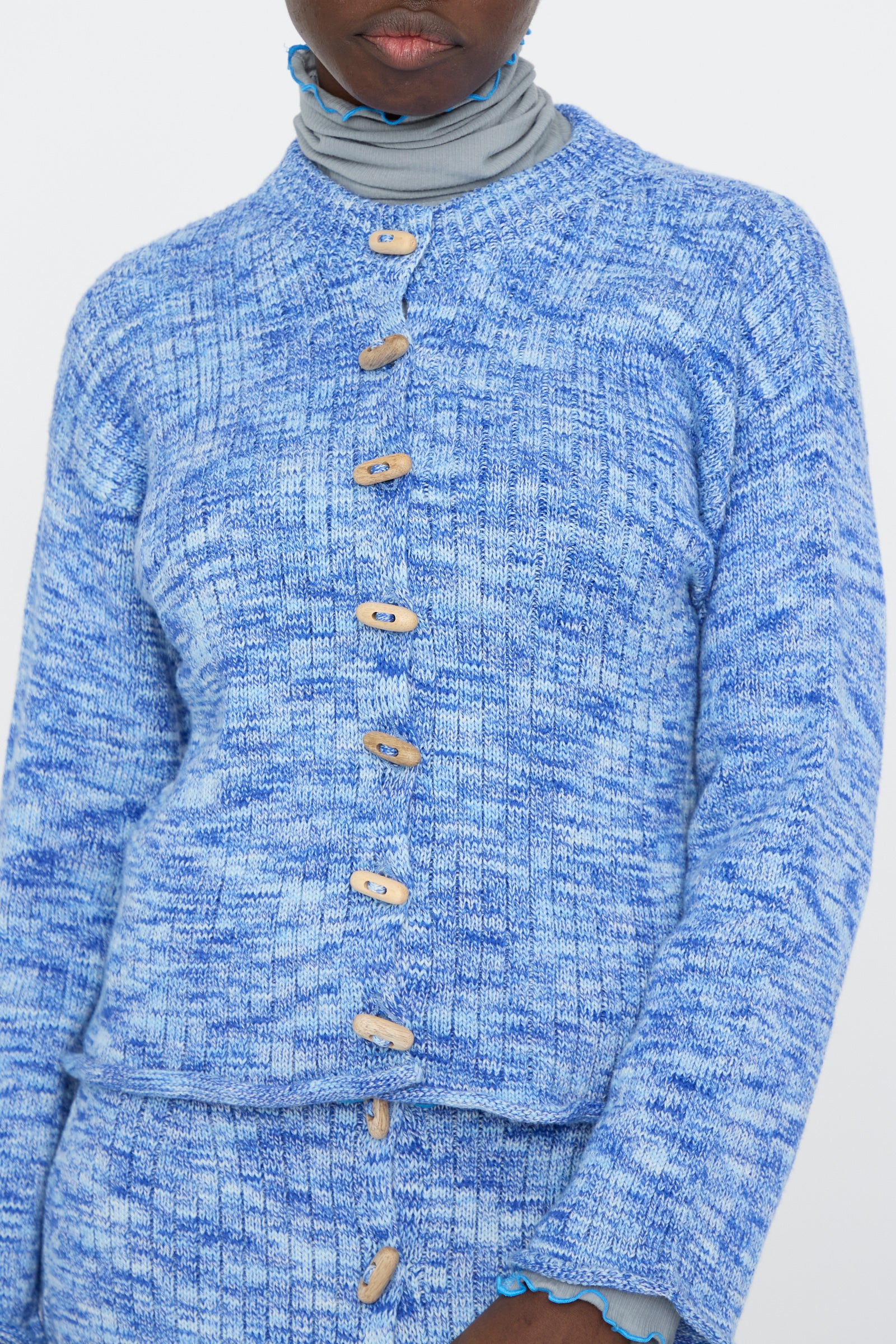 A person wears a Baserange Twisted Cotton Drip Cardigan in Blue Melange with wooden buttons over a gray turtleneck against a white background. 