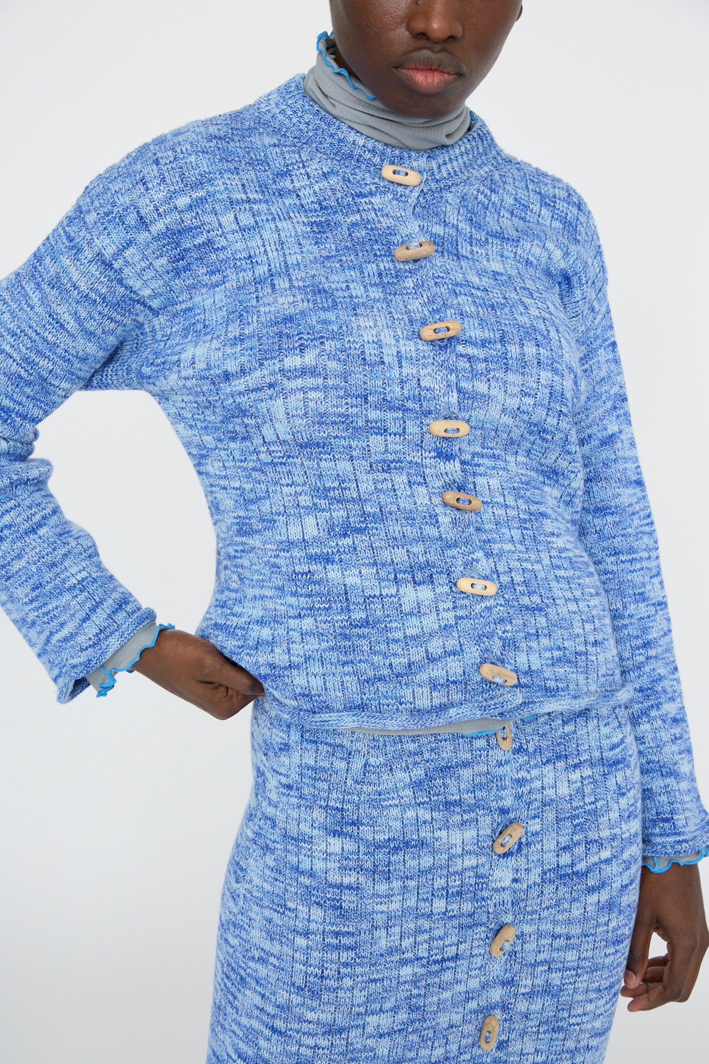 A person wearing a Baserange Twisted Cotton Drip Cardigan in Blue Melange paired with a skirt set, featuring elegant wooden buttons with an arm on their hip against a plain background. 