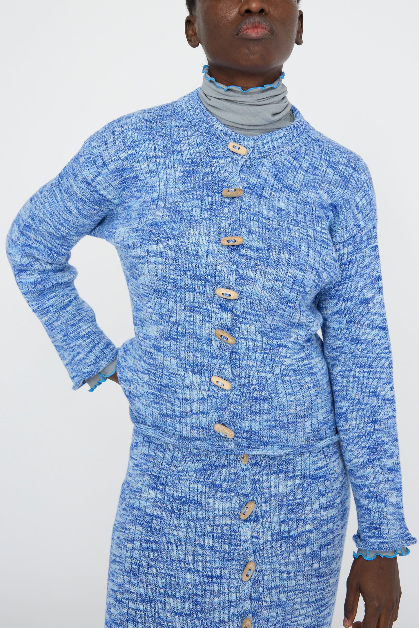 Wearing Baserange's Twisted Cotton Drip Cardigan in Blue Melange with wooden buttons, paired with a striped turtleneck and skirt, against a white backdrop.