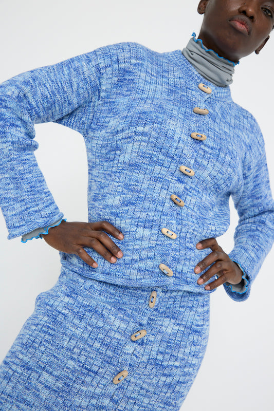 A person is wearing Baserange's Twisted Cotton Drip Cardigan in Blue Melange, paired with a matching skirt against a white backdrop. Both are crafted from organic cotton and feature round wooden buttons.